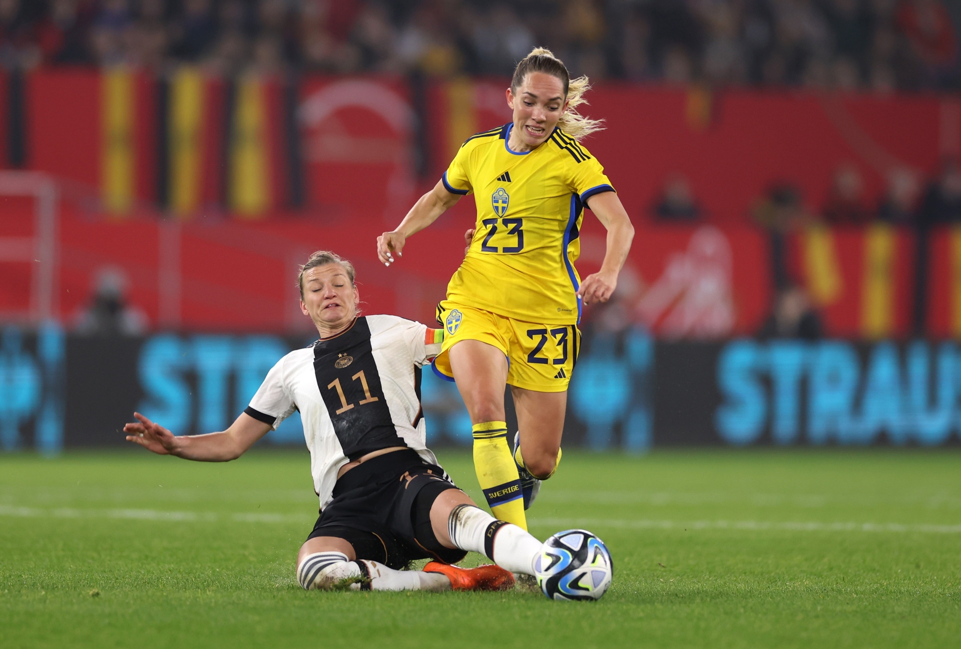 [{"clang_id":1,"value":"Zwei Fußballspielerinnen im Wettkampf. Eine Spielerin mit blonden Haaren in gelbem Trikot und Nummer 23 läuft. Die andere Spielerin in schwarz-weiß gestreiftem Trikot mit Nummer 11 rutscht auf dem Rasen. Im Hintergrund Stadion."}]