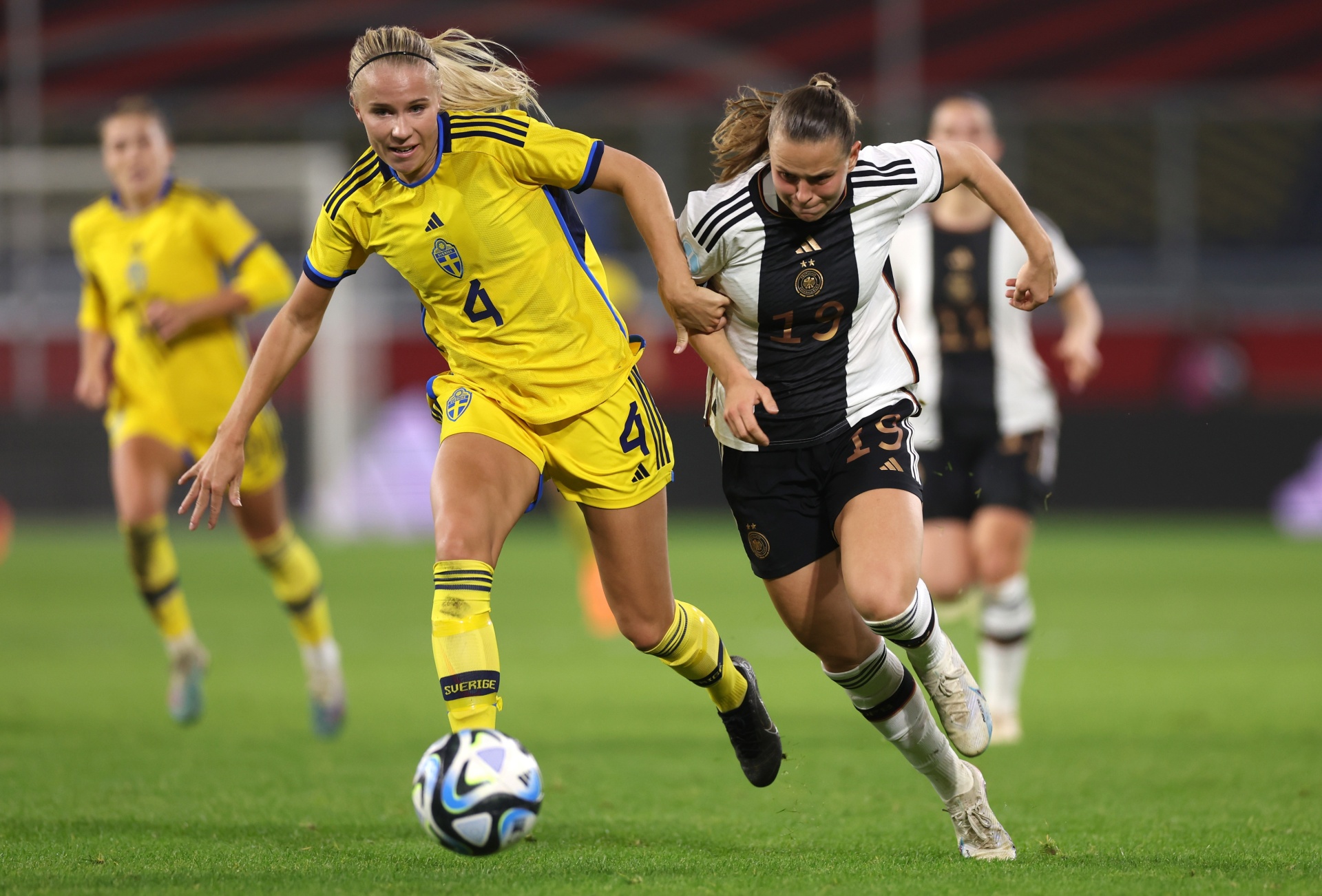 [{"clang_id":1,"value":"Zwei Fußballspielerinnen während eines Spiels: Links in gelben Trikots mit der Nummer 4, rechts in schwarz-weiß gestreiften Trikots mit der Nummer 15. Sie dribbeln den Ball auf einem Fußballfeld."}]