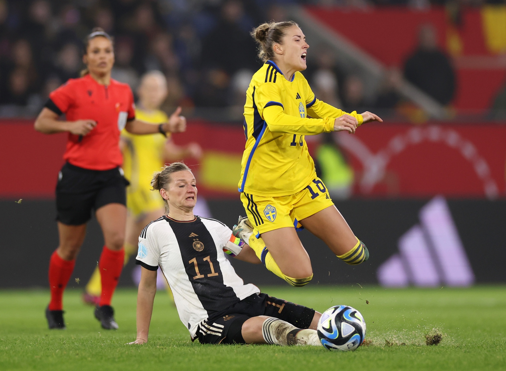 [{"clang_id":1,"value":"Zwei Fußballspielerinnen im Spiel: Eine in gelbem Trikot springt über eine in schwarz-weißem Trikot. Im Hintergrund eine Schiedsrichterin in rot. Rasenplatz."}]