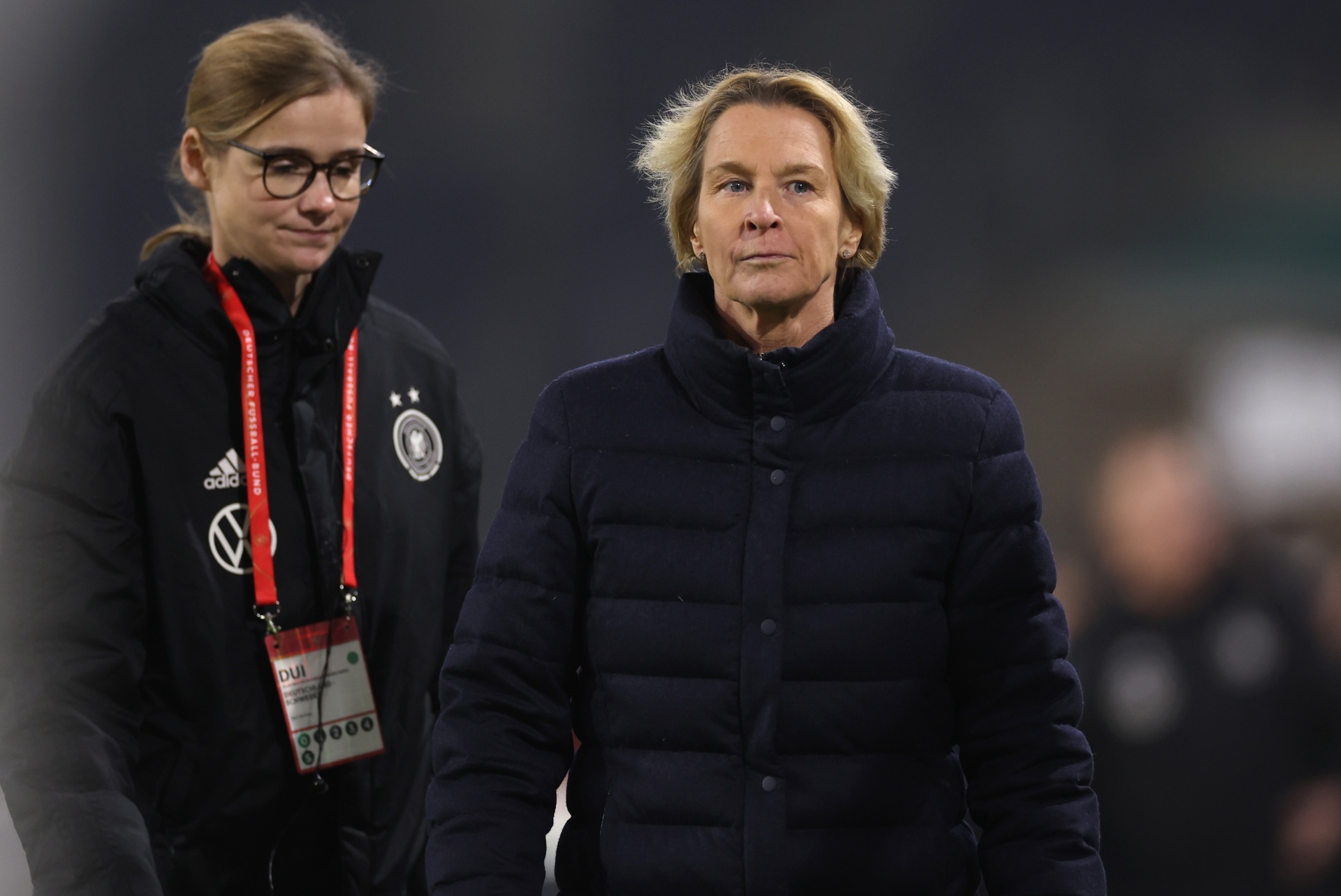 [{"clang_id":1,"value":"Zwei Frauen mit Sportbekleidung auf einem Fußballfeld. Eine trägt eine dunkle Jacke mit Adidas-Logo und Brille, die andere eine schwarze Jacke mit Sternen-Motiv."}]