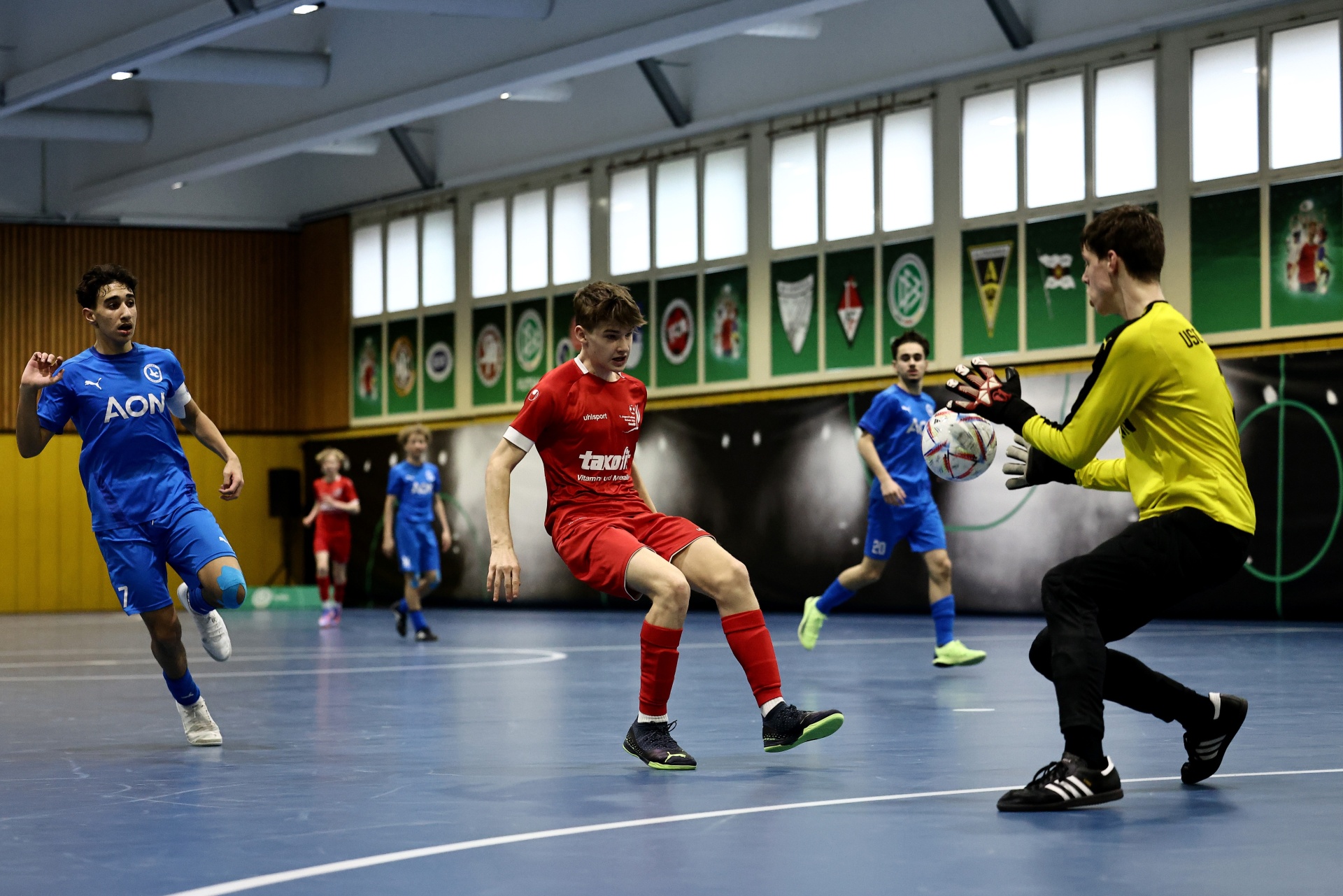 [{"clang_id":1,"value":"Futsalspiel in Sporthalle: Spieler in Rot schießt auf Torwart in Gelb, blauer Spieler im Hintergrund."}]