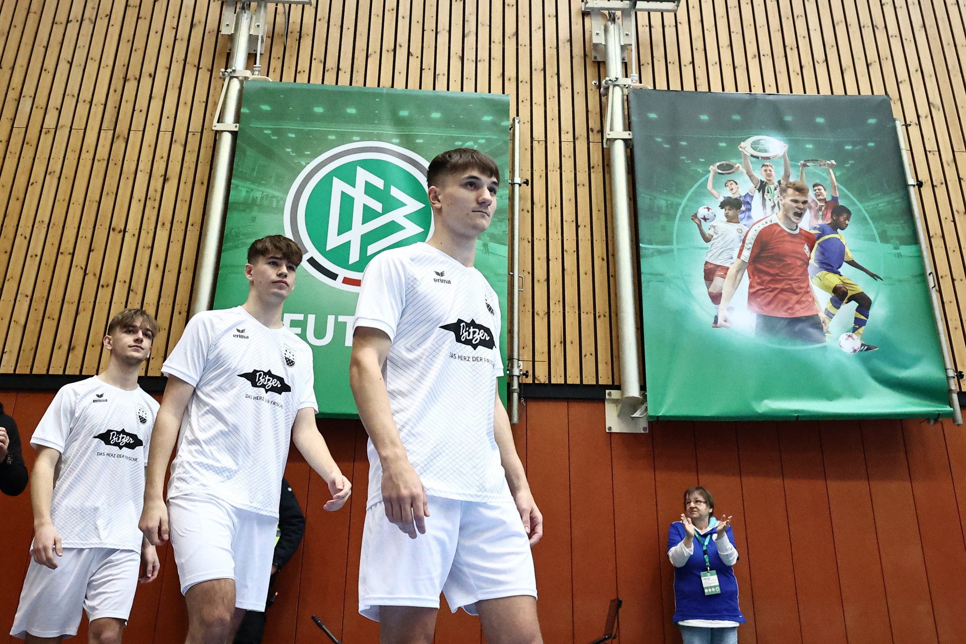 [{"clang_id":1,"value":"Drei junge Männer in weißen Sporttrikots und -shorts stehen auf einer Bühne vor einer Werbebannerwand mit Fußballmotiven. Rechts eine Frau mit Applaus."}]