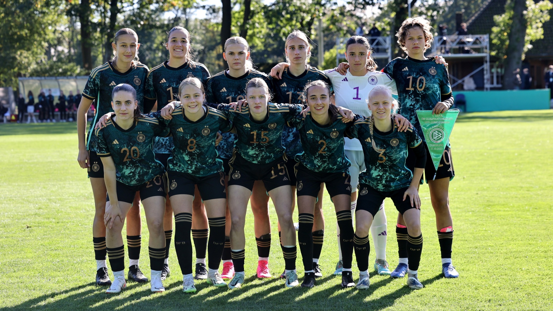 [{"clang_id":1,"value":"Fußballmannschaft, Frauen, in grünen Trikots und schwarzen Shorts auf Rasen. Spielerinnen stehen nebeneinander, lächeln, teilweise mit Nummern auf Trikots. Im Hintergrund Bäume und Gebäude."}]