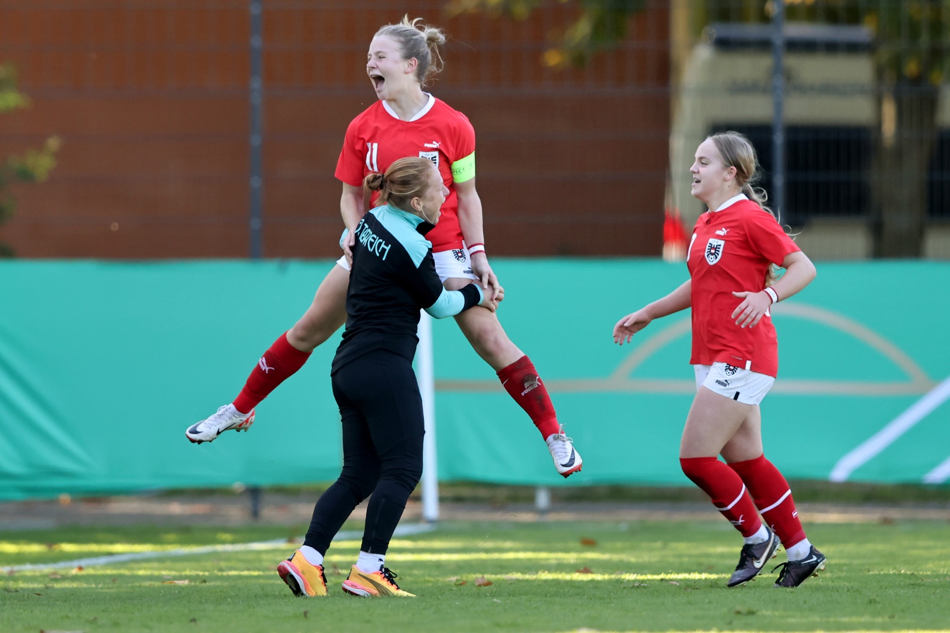[{"clang_id":1,"value":"Drei Fußballspielerinnen in roten Trikots und eine in dunklem Trikot feiern auf einem grünen Rasen. Eine Spielerin wird hochgehoben. Im Hintergrund ein Zaun und Bäume."}]