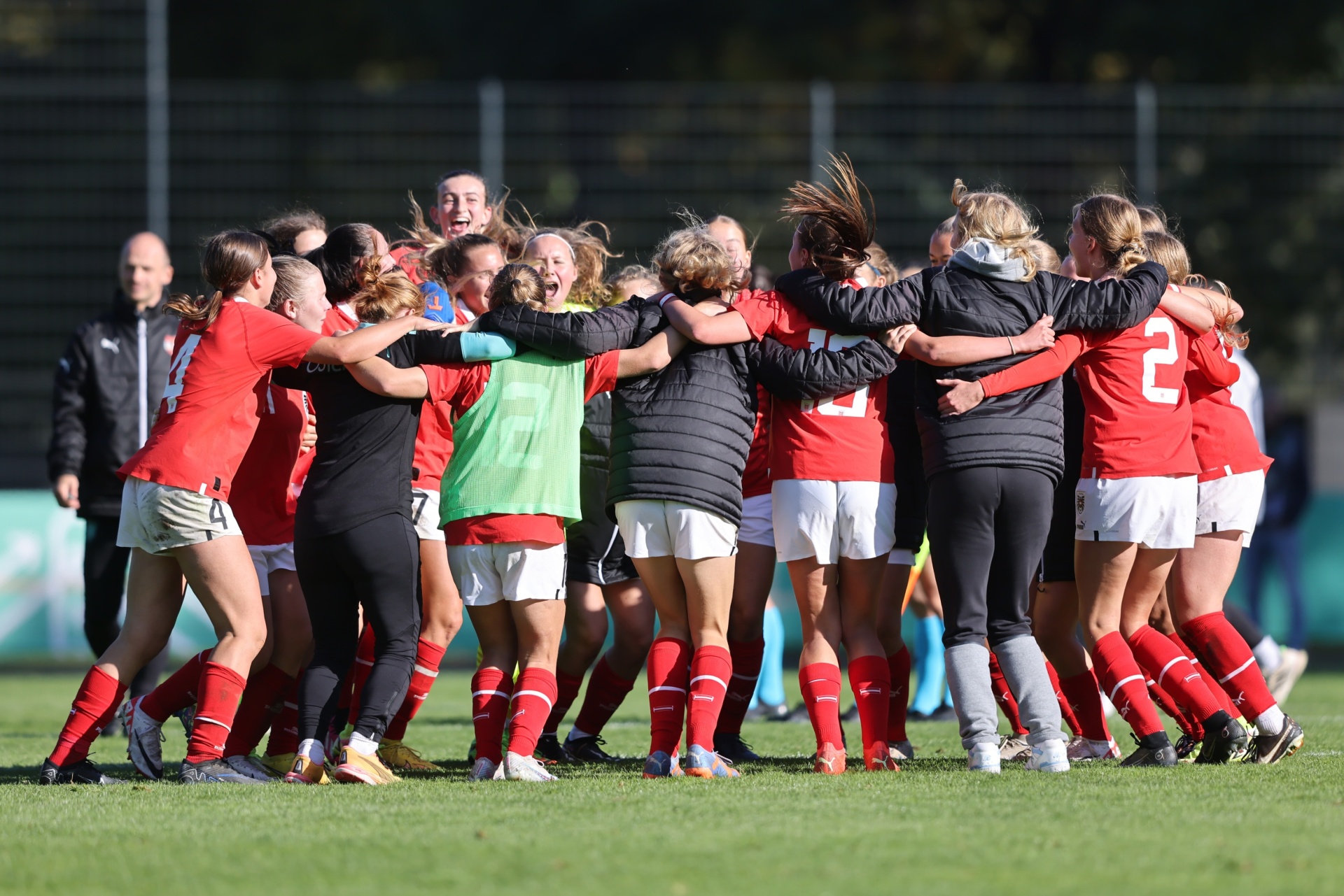 [{"clang_id":1,"value":"Frauenfußballteam im roten Trikot feiert eng umschlungen auf dem Rasen. Im Hintergrund steht ein Mann und eine Metallwand."}]