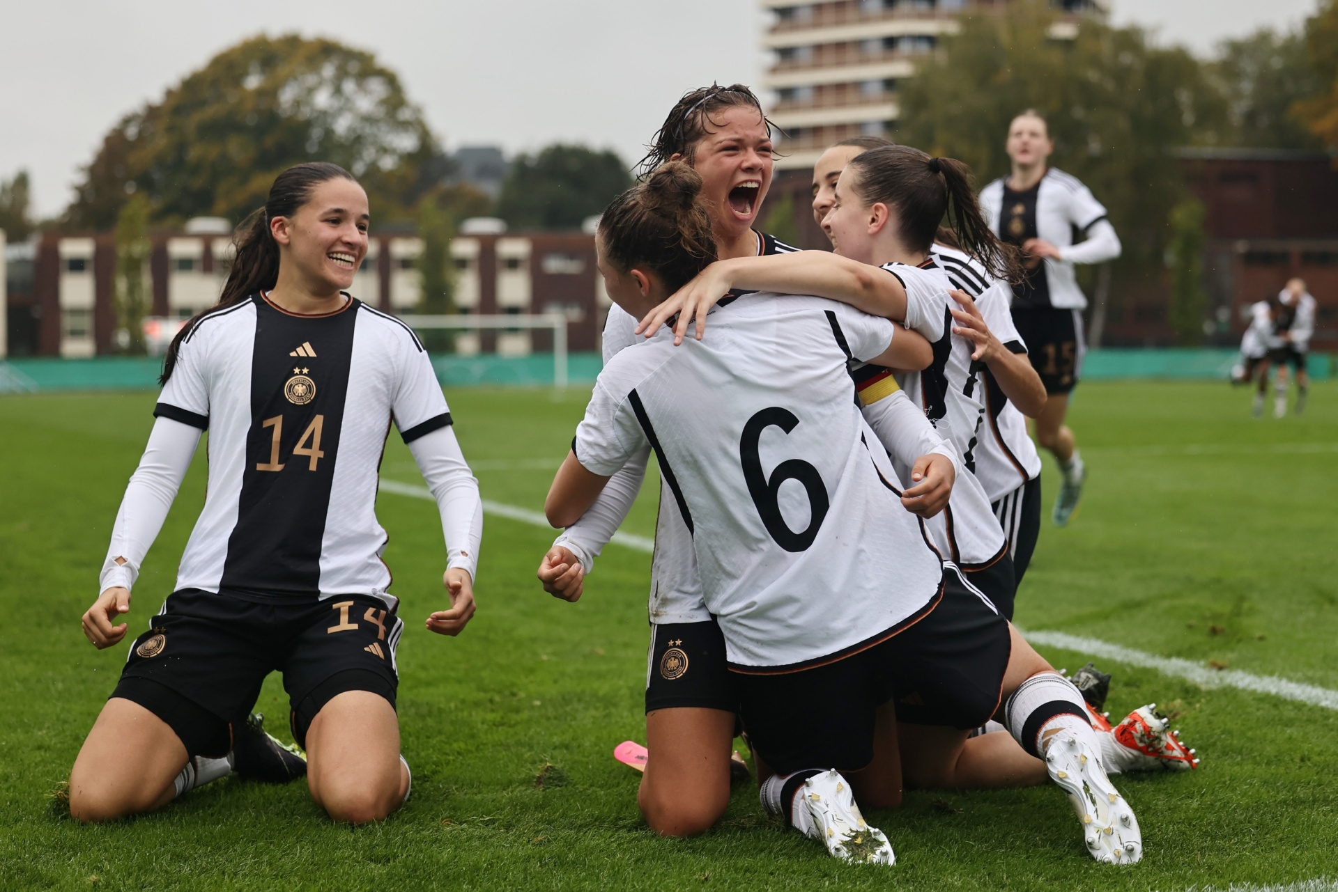 [{"clang_id":1,"value":"Frauenfußballteam Deutschland jubelt auf grünem Rasen. Vier Spielerinnen umarmen sich, Nummern 6 und 14 erkennbar. Im Hintergrund Gebäude und weitere Spielerinnen."}]