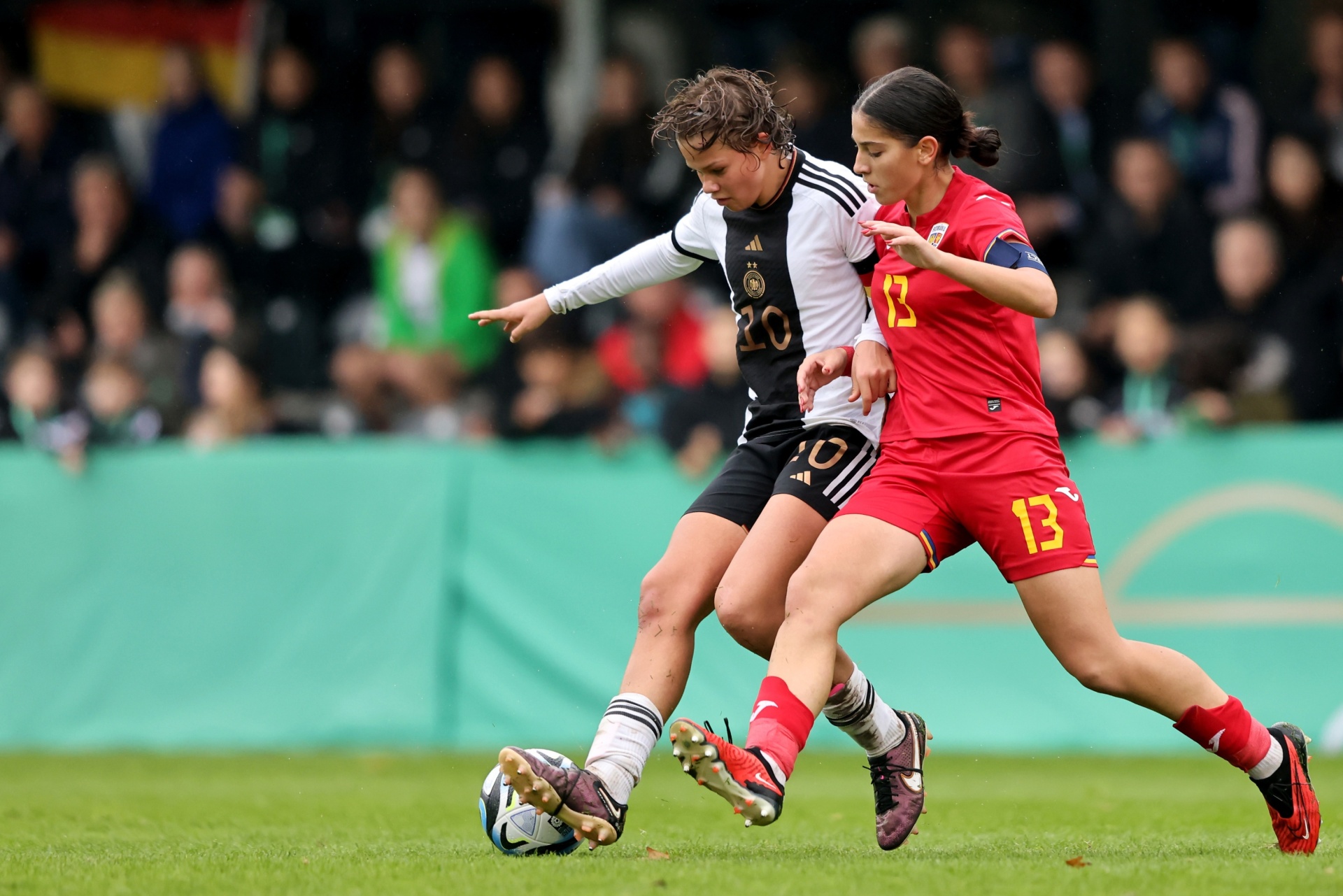 [{"clang_id":1,"value":"Zwei Fußballspielerinnen im Spiel. Links in Schwarz und Weiß, rechts in Rot, kämpfen um den Ball auf Grün. Im Hintergrund Zuschauer."}]