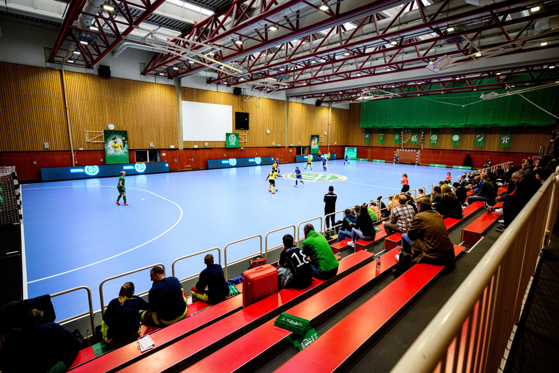 [{"clang_id":1,"value":"Futsal-Halle mit Spielern auf blauem Feld. Rote Zuschauertribüne mit sitzenden Zuschauern. Holzverkleidete Wände, Metallgebälk. Großbildschirm zeigt Spielszene."}]