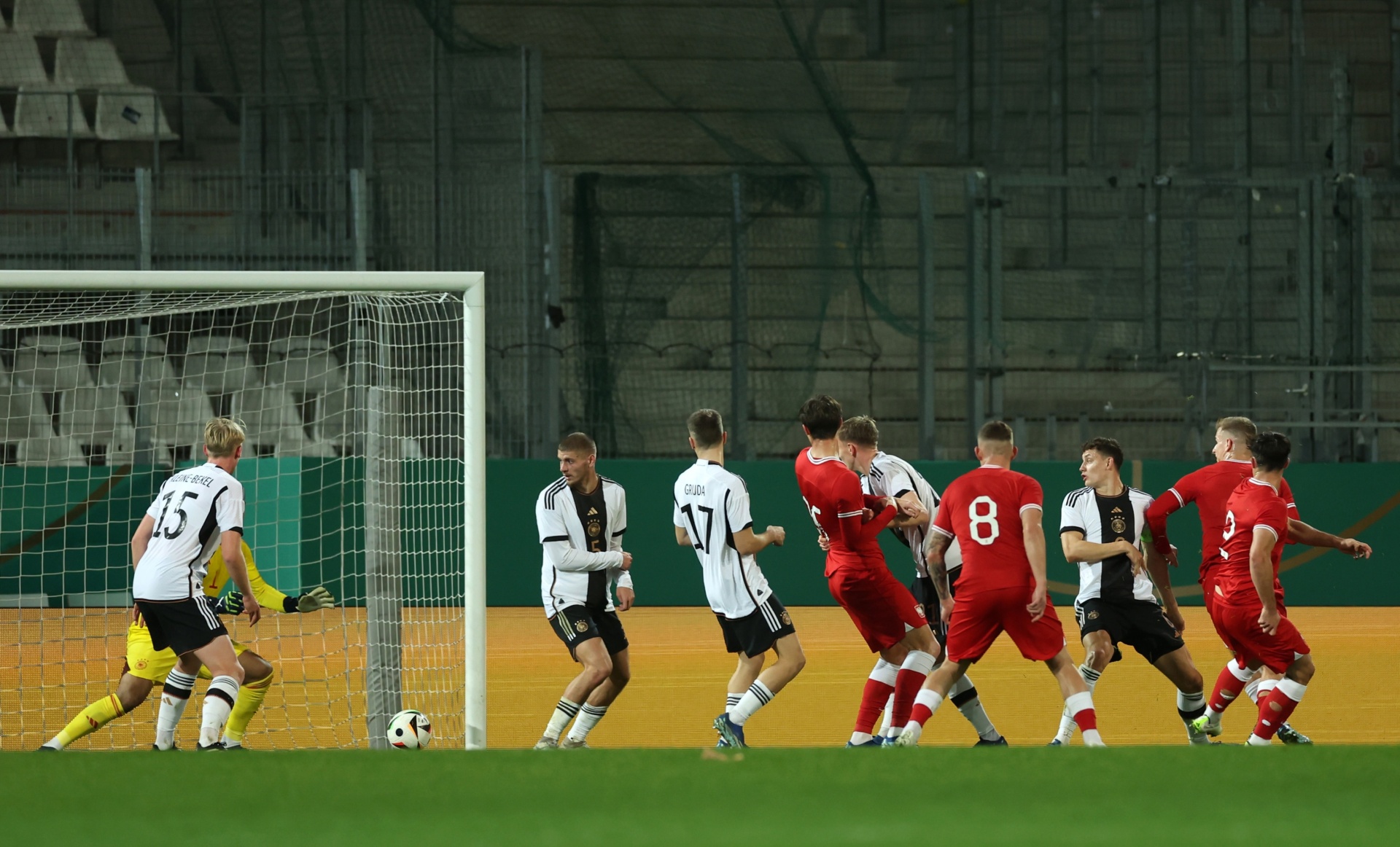 [{"clang_id":1,"value":"Fußballspiel: Zwei Mannschaften stehen sich gegenüber. Links Torwart, rechts Spieler in roten Trikots und schwarzer Hose/Shorts. Rasenplatz, Stadion im Hintergrund."}]