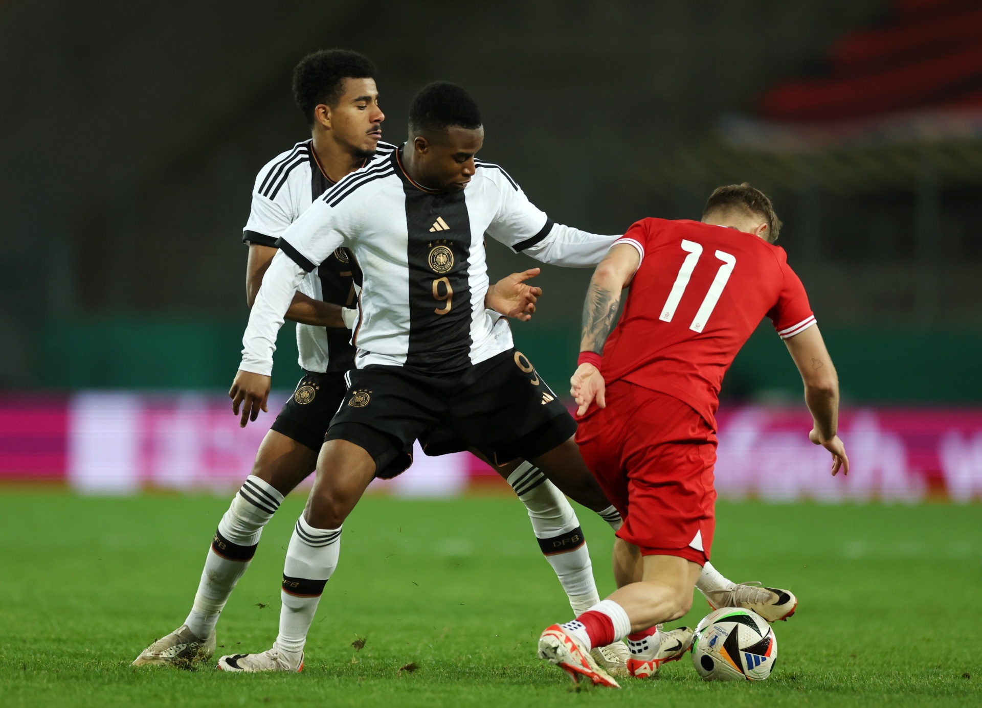 [{"clang_id":1,"value":"Fußballspiel: Zwei Spieler mit Deutschland-Trikot (9 und unbekannte Nummer) kämpfen um den Ball gegen einen Spieler mit rotem Trikot (11) auf Grün."}]