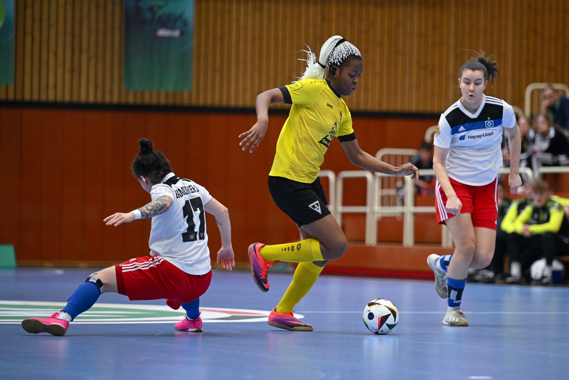 [{"clang_id":1,"value":"Futsal-Spiel: Spielerinnen in Gelb, Rot/Schwarz und Weiß/Rot kämpfen um den Ball auf blauem Hallenboden. Eine Spielerin dribbelt, eine rutscht zum Blocken."}]