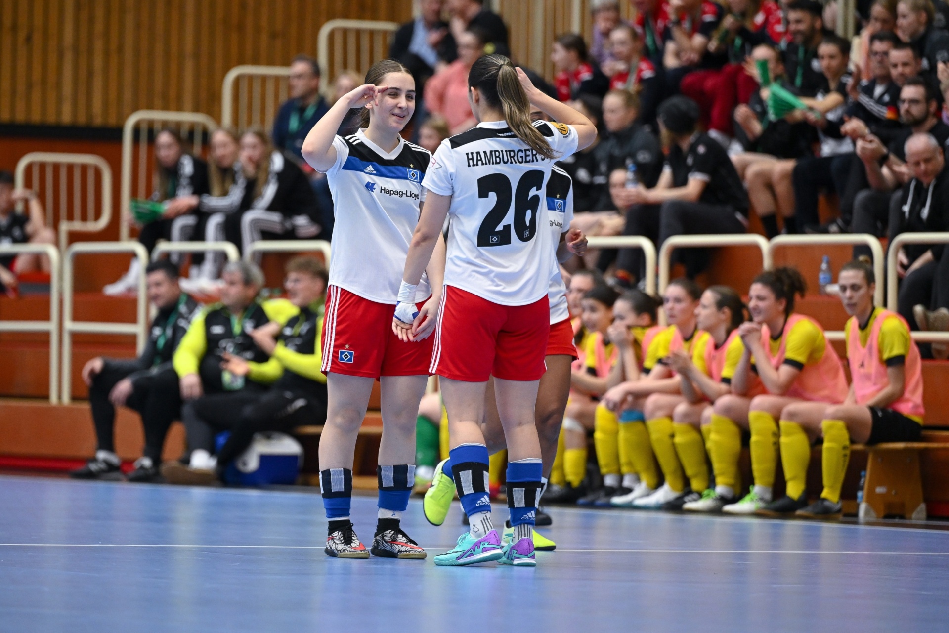 [{"clang_id":1,"value":"Zwei Handballspielerinnen feiern auf dem Spielfeld. Im Hintergrund sitzen oder stehen Zuschauende in einer Sporthalle."}]