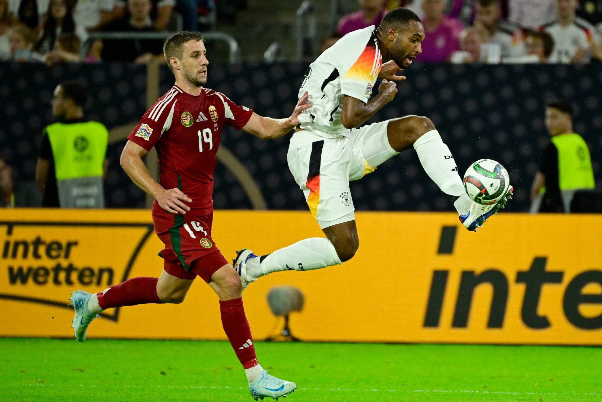[{"clang_id":1,"value":"Zwei Fußballspieler im Spiel. Links ein Spieler mit rotem Trikot (Nummer 19), rechts ein Spieler in Weiß-Schwarz mit dem Ball am Fuß, in Luft akrobatisch. Grünrasen, Stadionhintergrund."}]