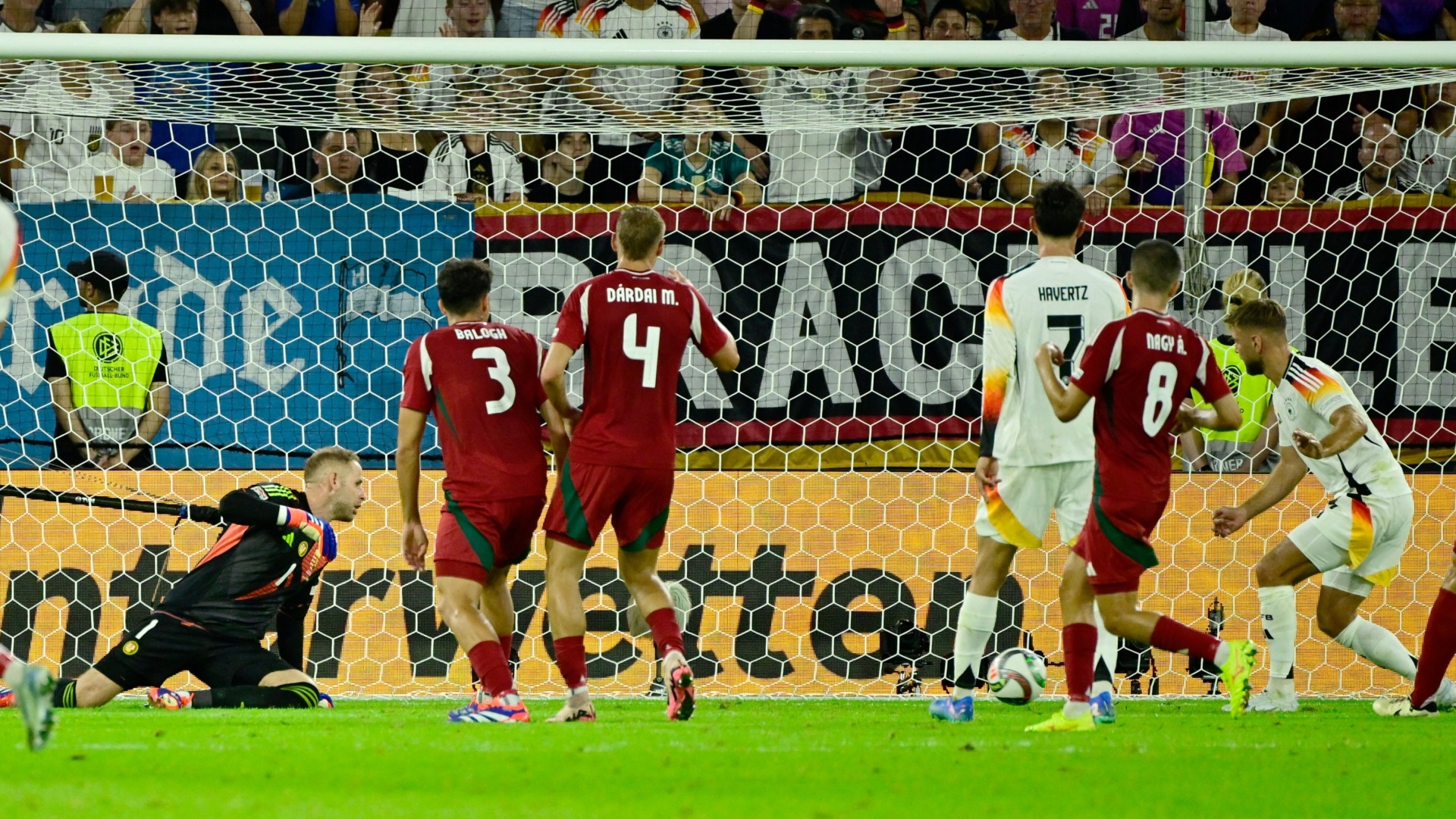 [{"clang_id":1,"value":"Fußballspiel: Torwart springt, um Schuss zu halten. Spieler in roten und weißen Trikots stehen im Strafraum. Hintergrund: Zuschauer und Werbebanner."}]