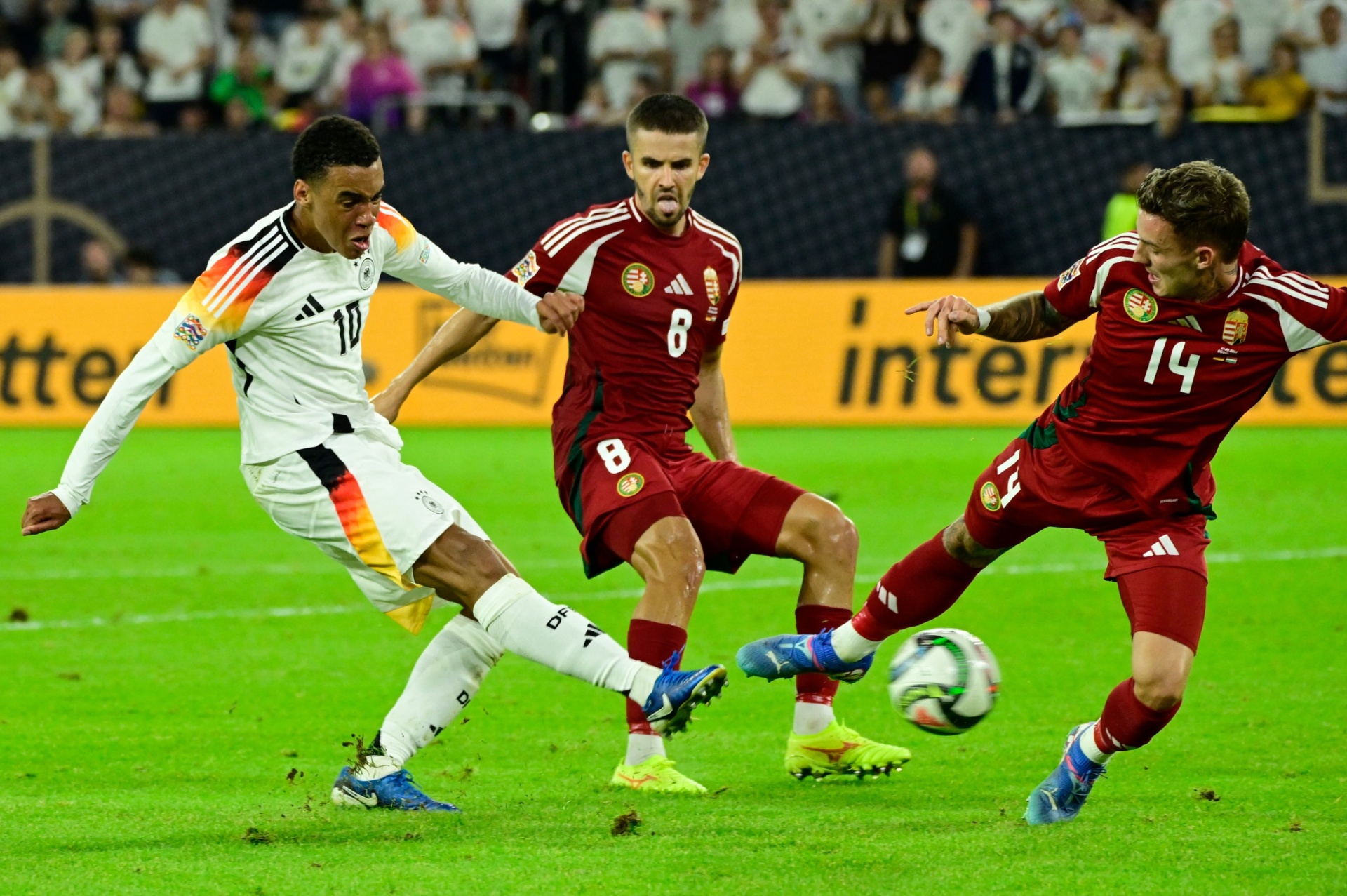 [{"clang_id":1,"value":"Fußballspiel: Spieler mit Trikotnummer 10 (weiß) schießt Ball, zwei Gegenspieler (rot) versuchen Blocken. Rasenplatz, Stadion im Hintergrund."}]
