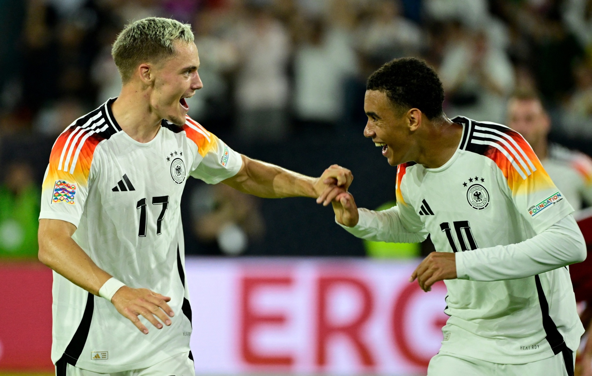 [{"clang_id":1,"value":"Zwei Fußballspieler in weißen Trikots mit deutschen Flaggenfarben jubeln vor einem Stadion. Spieler 17 und 10, Blick nach rechts. Im Hintergrund „ERC“ in roten Buchstaben."}]
