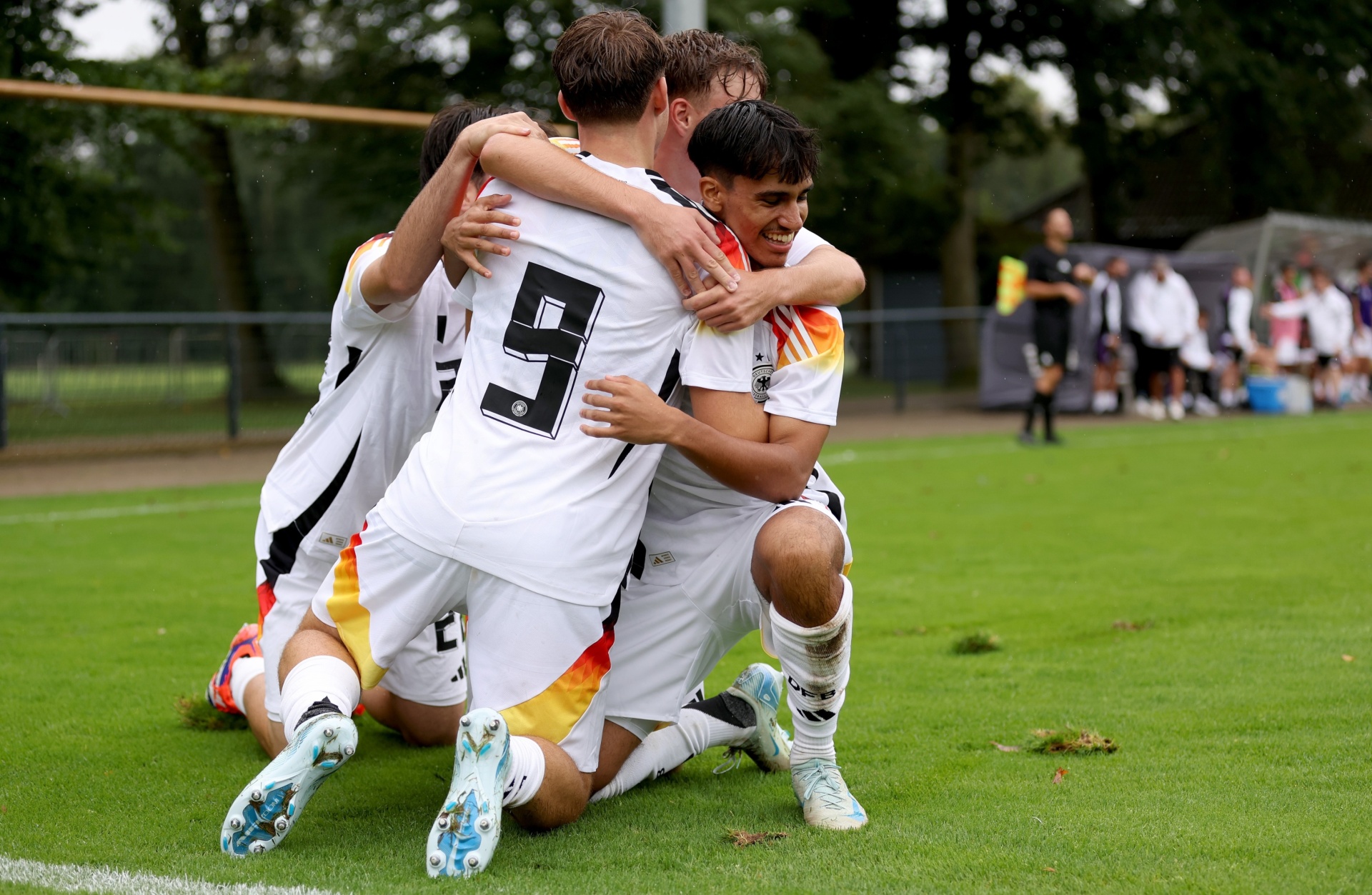 [{"clang_id":1,"value":"Fußballspieler in weißen Trikots mit den deutschen Farben feiern einen Treffer. Spieler umarmen sich auf dem grünen Rasen. Im Hintergrund Zuschauer und Bäume."}]
