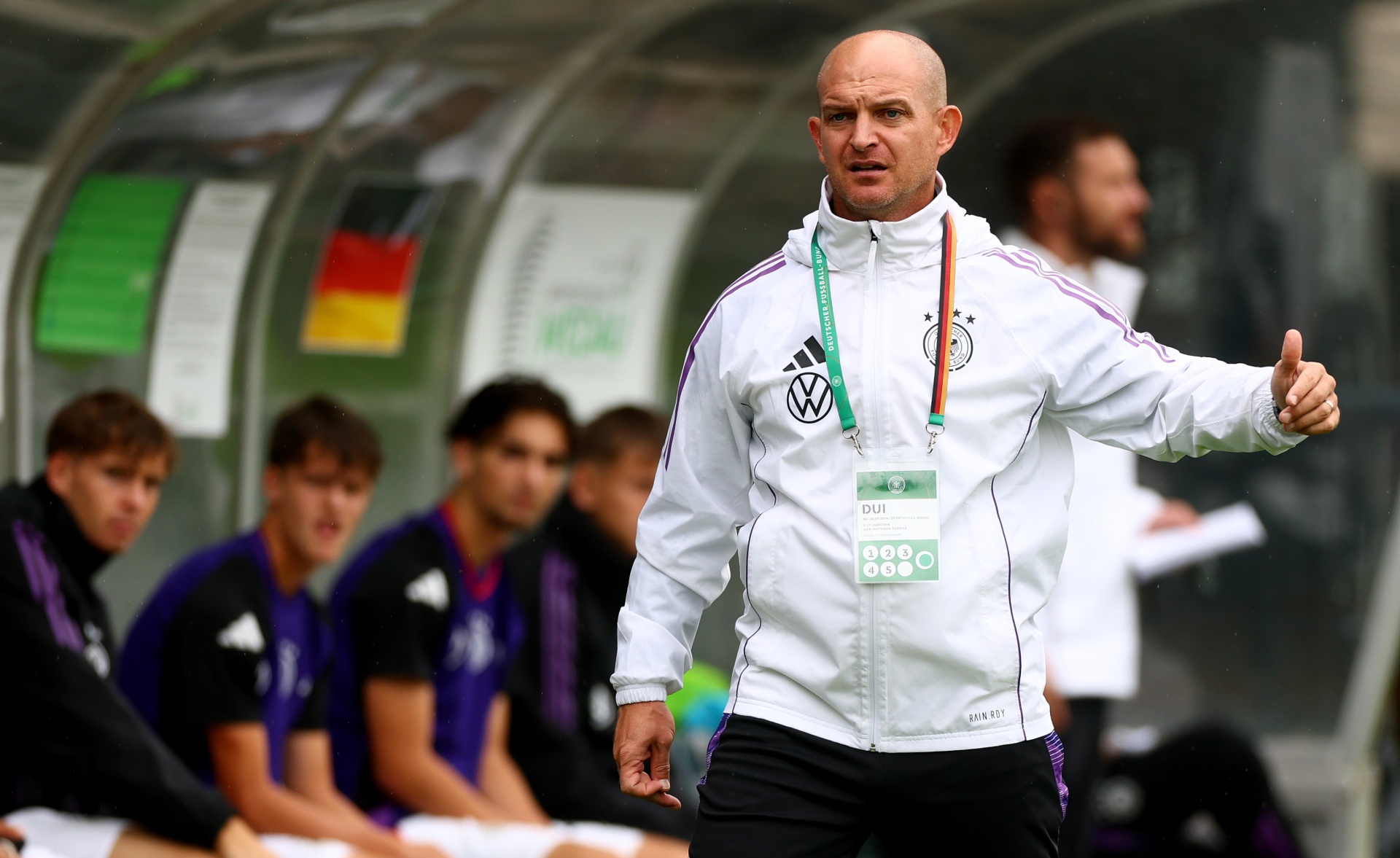 [{"clang_id":1,"value":"Fußballtrainer mit Glatze, weißer Trainingsjacke und Deutschland-Lanyard gibt Handzeichen. Im Hintergrund verschwommen Spieler auf der Bank."}]