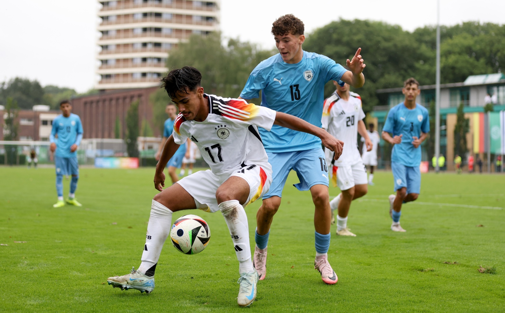 [{"clang_id":1,"value":"Zwei Fußballspieler im Spiel: Spieler mit Nummer 17 (Deutschland) dribbelt den Ball, wird von Spieler mit Nummer 13 (hellblaues Trikot) gedeckt. Rasen, Gebäude im Hintergrund."}]