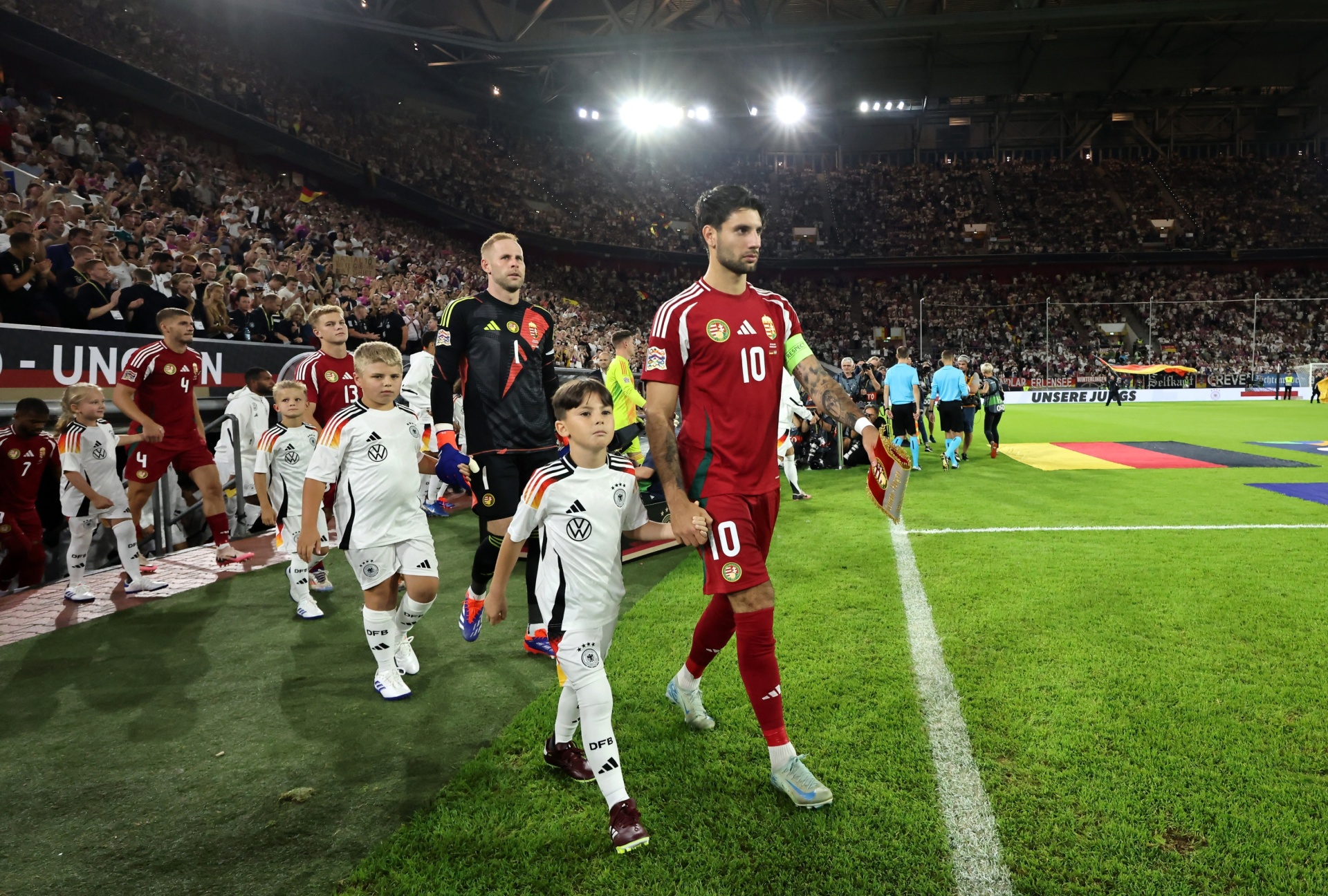 [{"clang_id":1,"value":"Fußballspieler in roten Trikots und Kindermasottchen im schwarzen und weißen Trikot betreten einen grünen Rasen vor gefülltem Stadion."}]