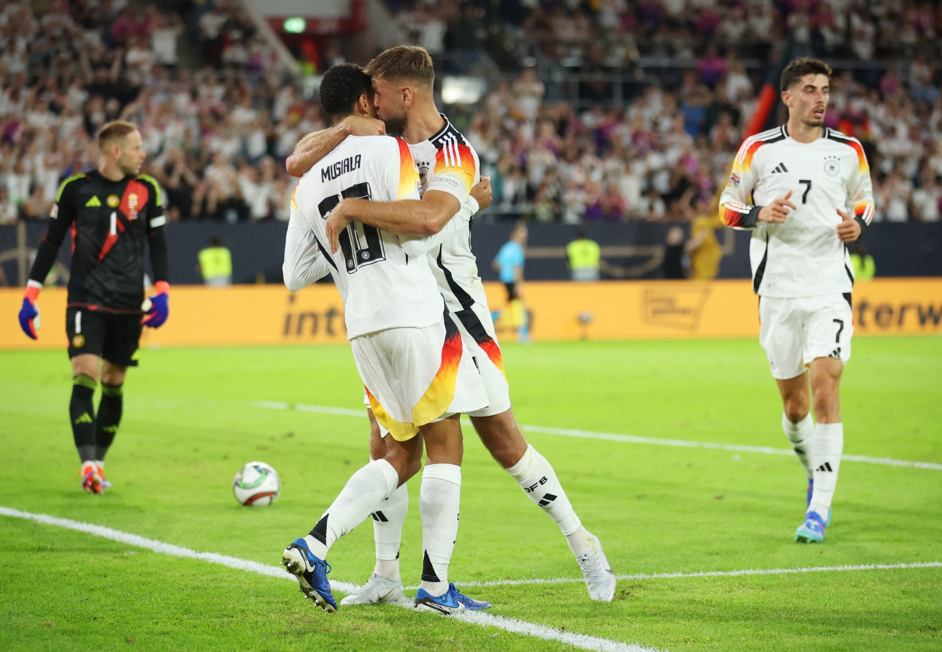 [{"clang_id":1,"value":"Zwei Fußballspieler in weißen Trikots mit deutschen Farben feiern ein Tor. Im Hintergrund sind weitere Spieler und Zuschauer auf einer grünen Rasenfläche zu sehen."}]