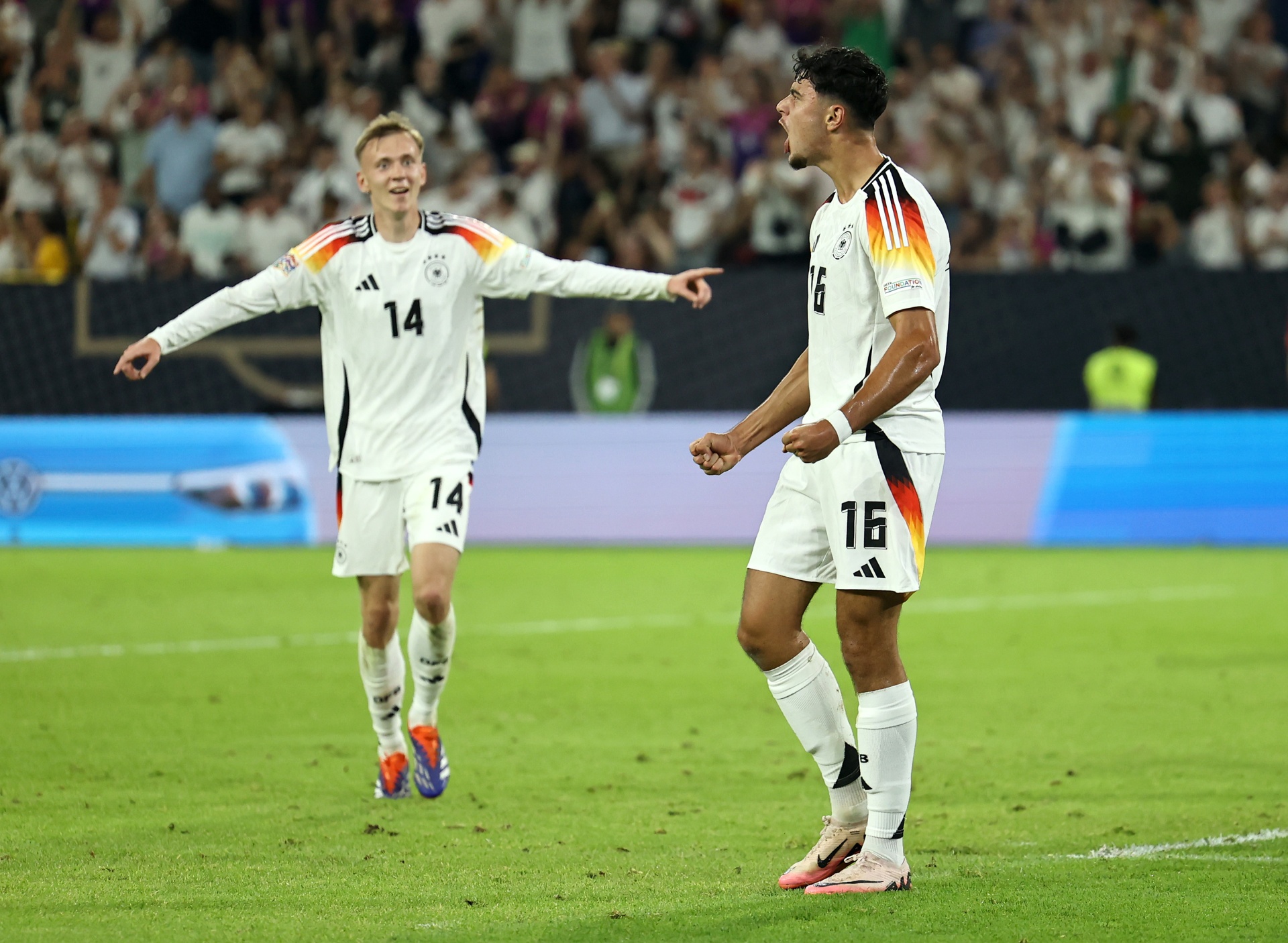 [{"clang_id":1,"value":"Zwei Fußballspieler in Deutschlandtrikots (Nummern 14 und 16) feiern auf einem grünen Rasen vor jubelndem Publikum."}]