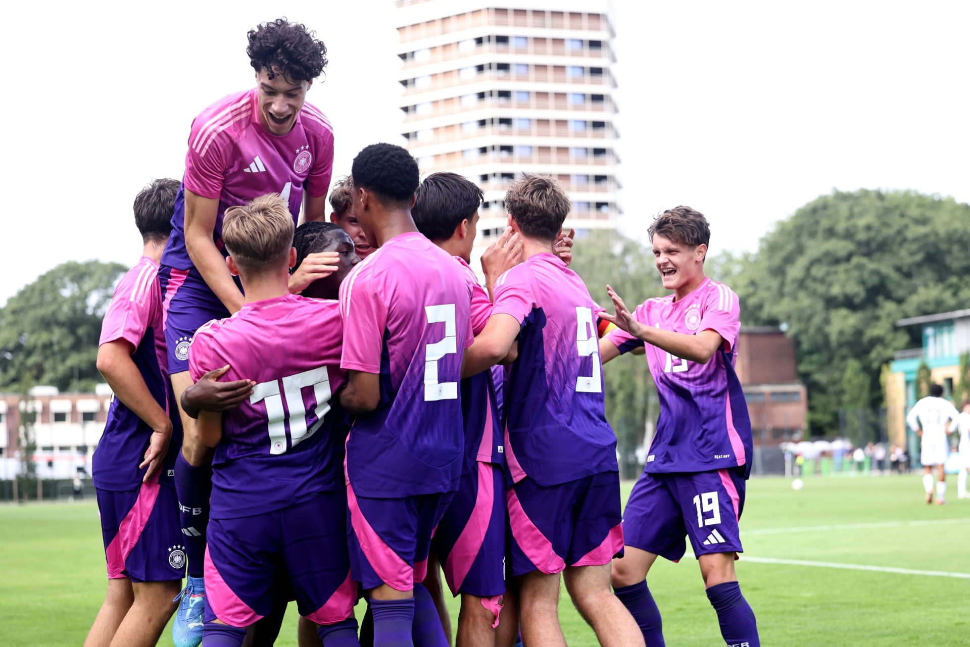 [{"clang_id":1,"value":"Fußballspieler in rosa und violetten Trikots feiern einen Treffer auf einem Rasenplatz. Im Hintergrund ein Wohngebäude und Bäume."}]