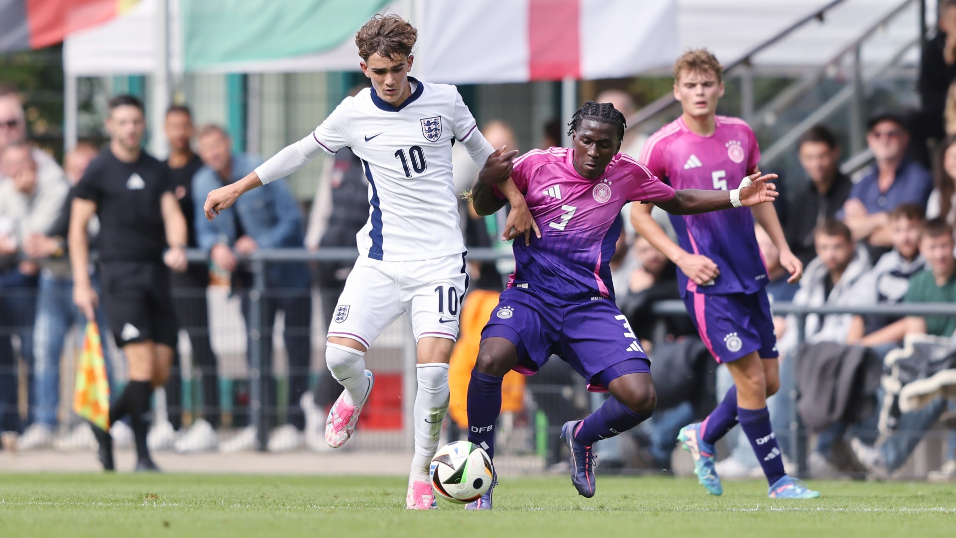 [{"clang_id":1,"value":"Zwei Fußballspieler im Spiel: Links Spieler mit weißem Trikot und Nummer 10, rechts Spieler mit violettem Trikot und Ball. Hintergrund: Zuschauer und Sportfeld."}]
