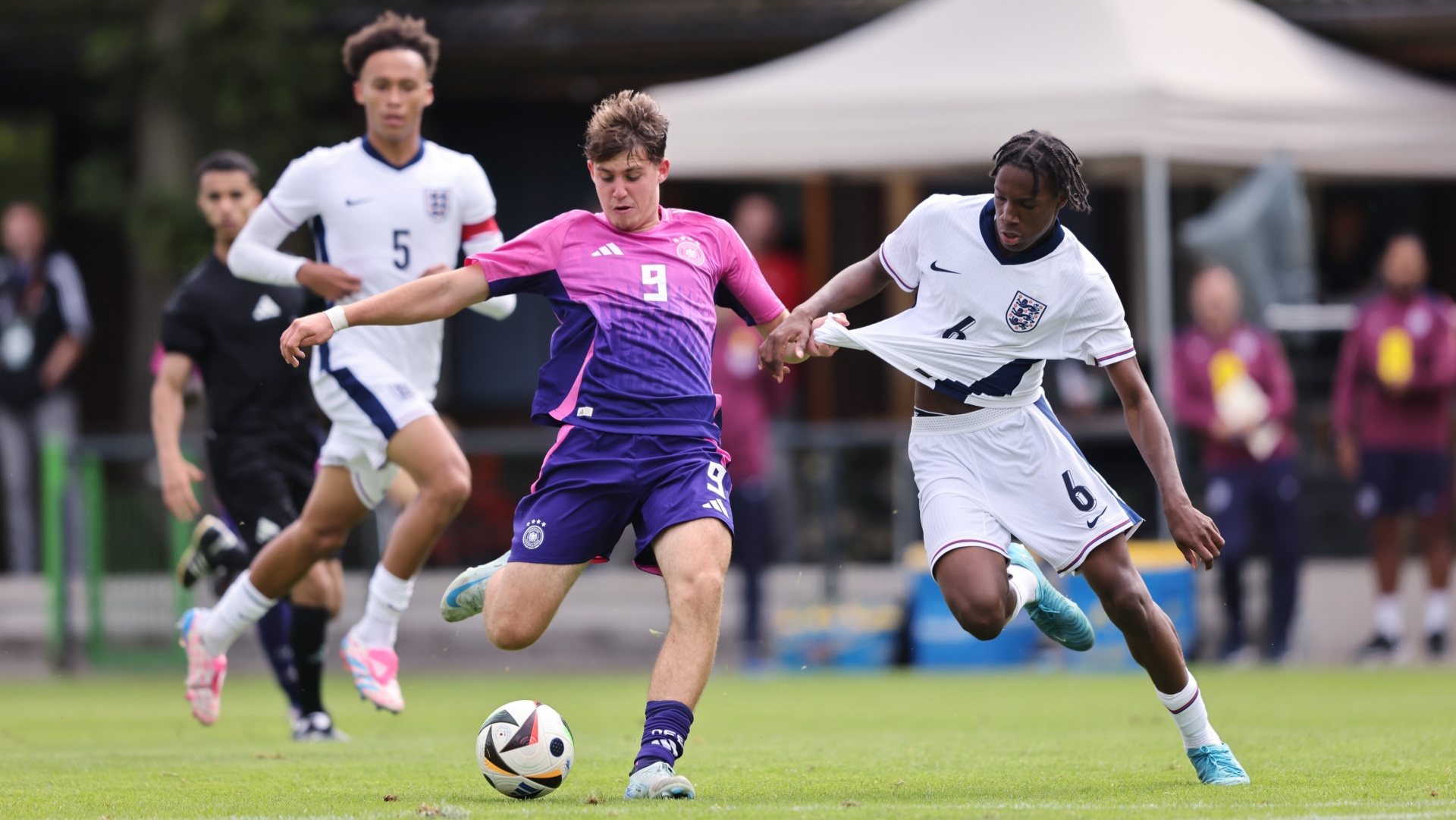 [{"clang_id":1,"value":"Fußballspiel: Spieler mit Nummer 9 (USA, lila Trikot) schießt Ball, wird von Spieler mit Nummer 6 (England, weiß-blaues Trikot) begleitet. Im Hintergrund weitere Spieler und Zuschauer."}]