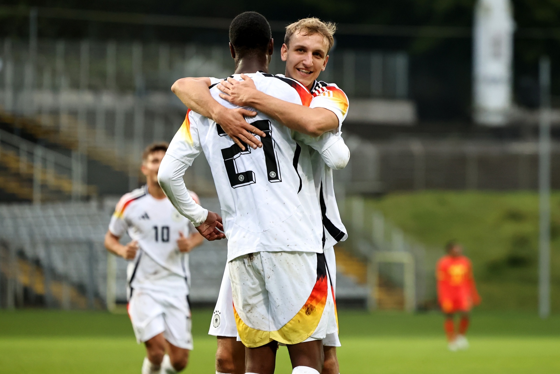 [{"clang_id":1,"value":"Zwei Fußballspieler im weißen Trikot mit deutschen Farben umarmen sich auf dem Rasen. Im Hintergrund weitere Spieler und Zuschauerränge."}]