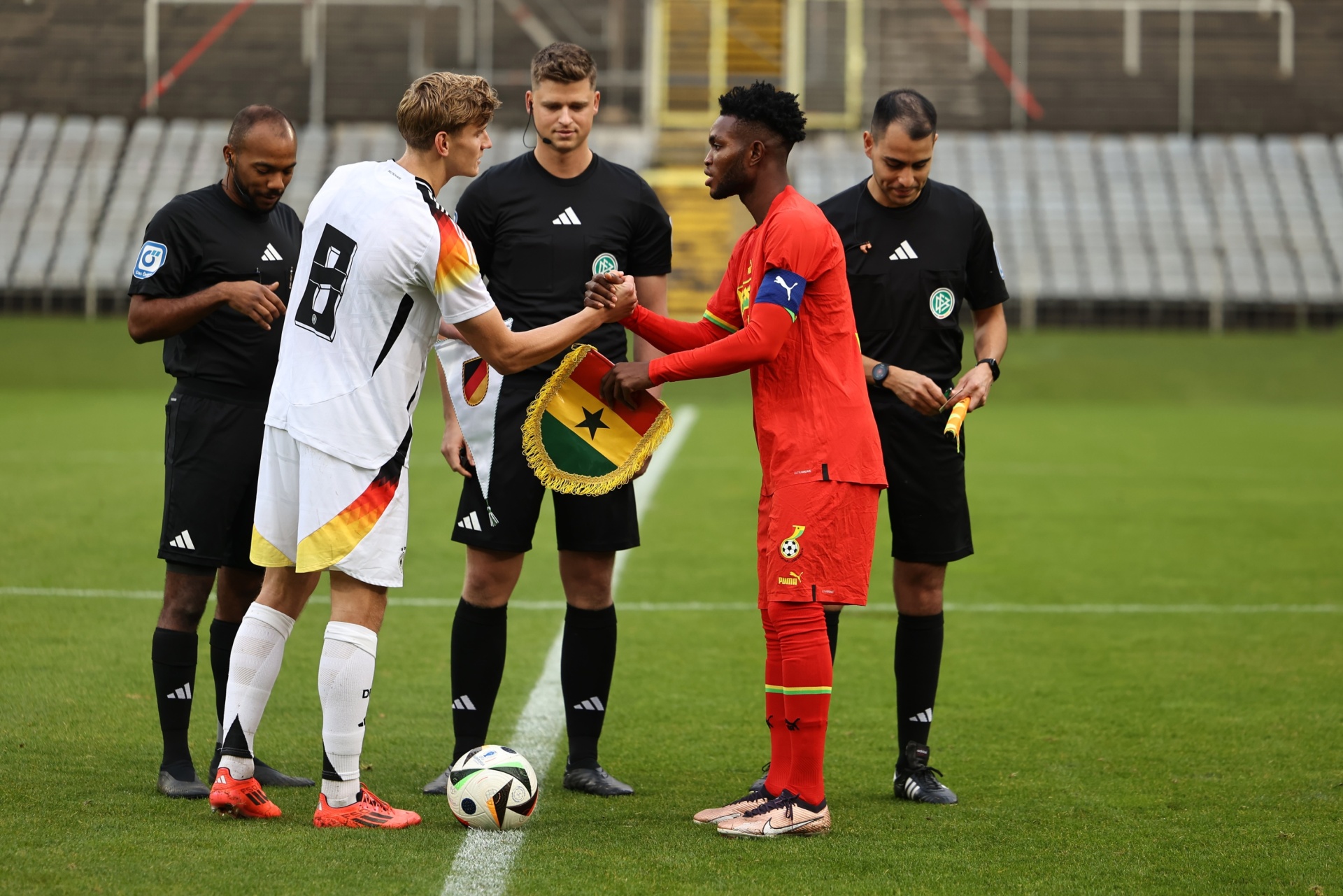 [{"clang_id":1,"value":"Zwei Fußballspieler, einer in Deutschland-Trikot, der andere in Ghana-Trikot, geben sich die Hand vor einem Spiel. Schiedsrichter stehen um sie herum, ein Fußball liegt auf dem Rasen."}]