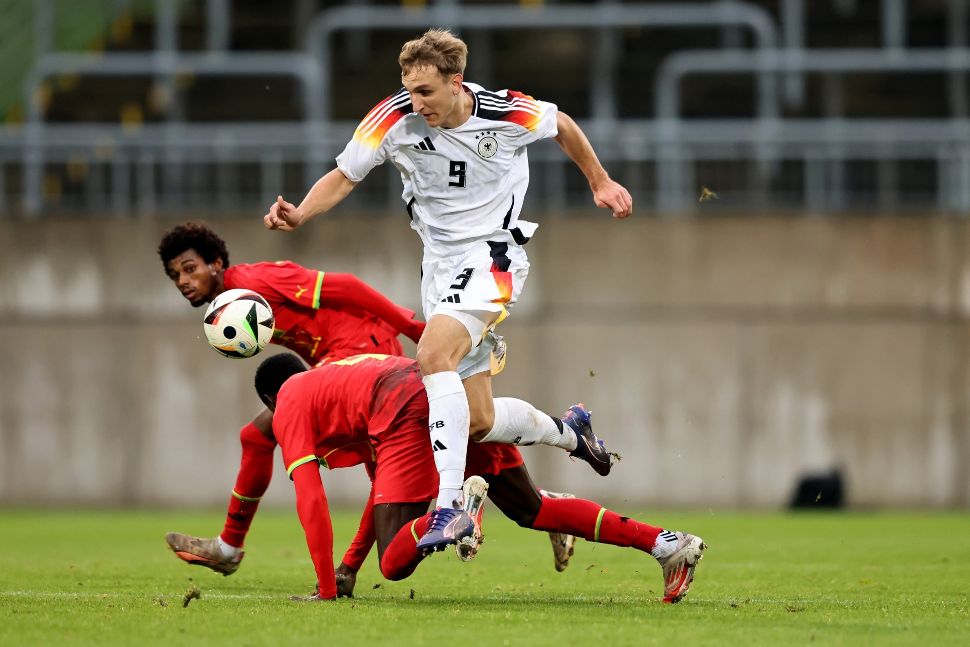 [{"clang_id":1,"value":"Fußballspiel: Spieler mit Trikotnummer 9 in Weiß/Schwarz dribbelt an zwei Gegenspielern in Rot vorbei. Rasenplatz und Zuschauerränge im Hintergrund."}]
