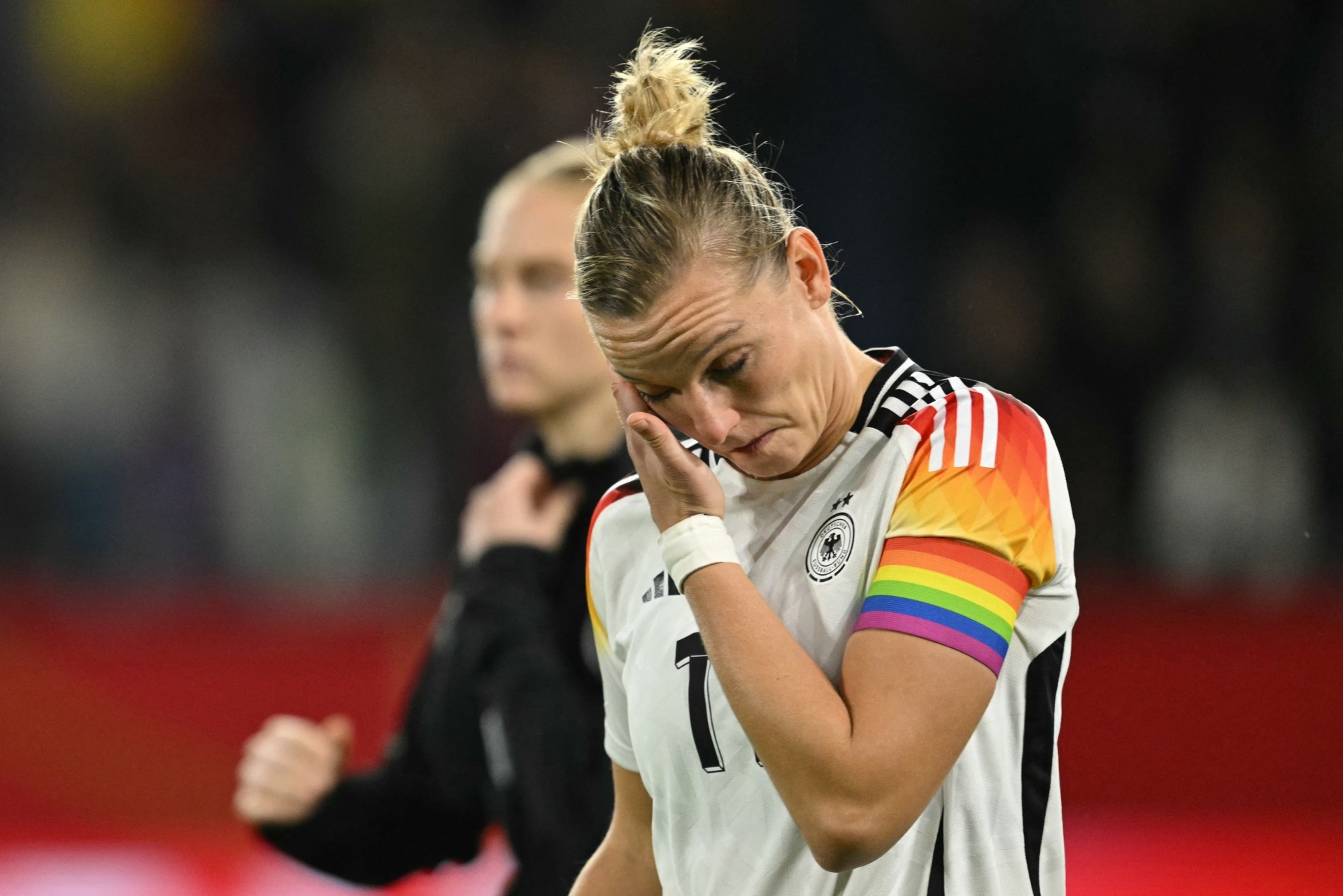[{"clang_id":1,"value":"Frau in Fußballtrikot mit Nummer 1, traurig, Kopf gesenkt. Ärmel mit Regenbogenfarben. Im Hintergrund verschwommene Personen und Stadion."}]