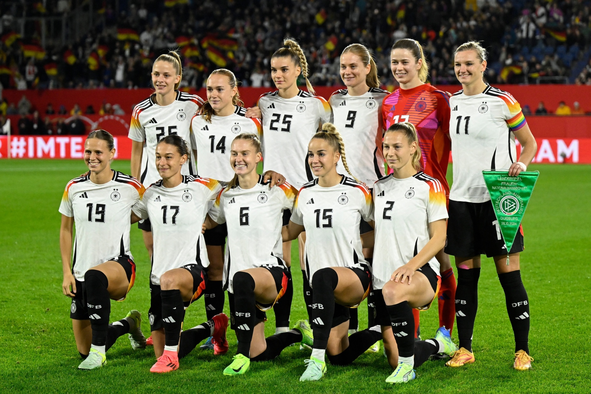 [{"clang_id":1,"value":"Frauenfußballmannschaft in weißen Trikots mit schwarzen Shorts und Stutzen. Sie posieren vor einem Stadion. Einige Spieler knien, andere stehen. Hintergrund verschwommen."}]