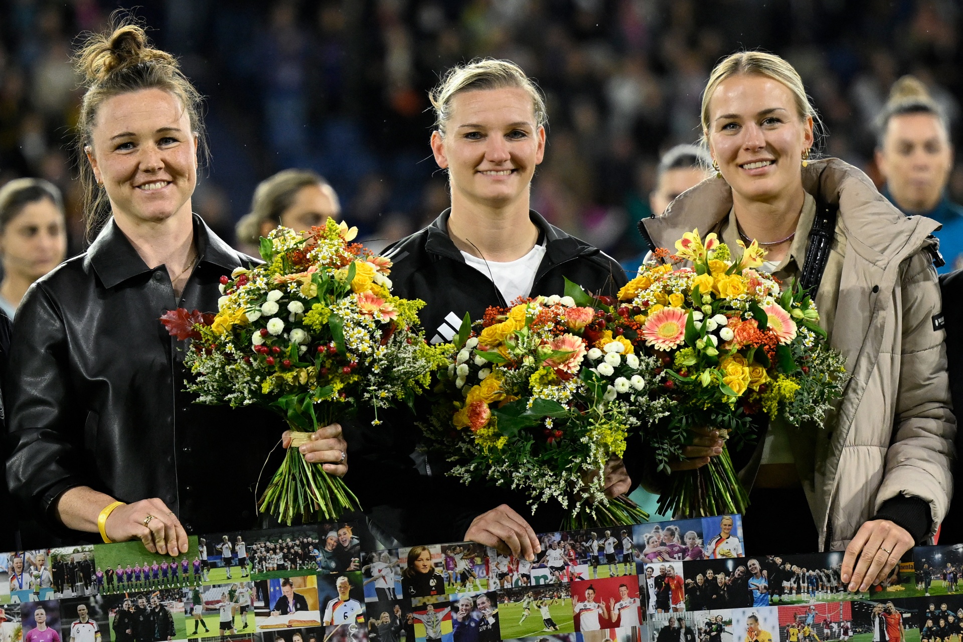 [{"clang_id":1,"value":"Drei lächelnde Fußballspielerinnen stehen nebeneinander, jede hält einen Blumenstrauß und einen Bilderbogen. Hintergrund: Zuschauer."}]