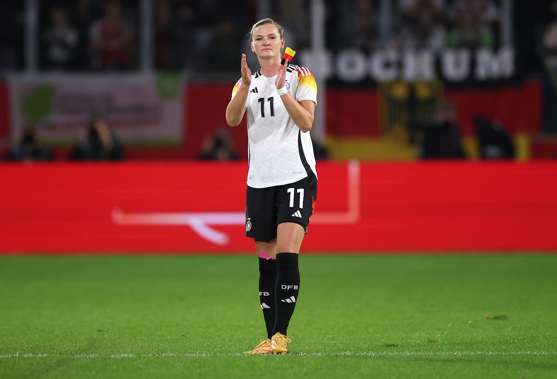 [{"clang_id":1,"value":"Fußballspielerin mit Trikotnummer 11 klatscht auf dem Rasen. Sie trägt schwarze Shorts, Stutzen und gelbe Fußballschuhe. Im Hintergrund Zuschauer und Stadion."}]