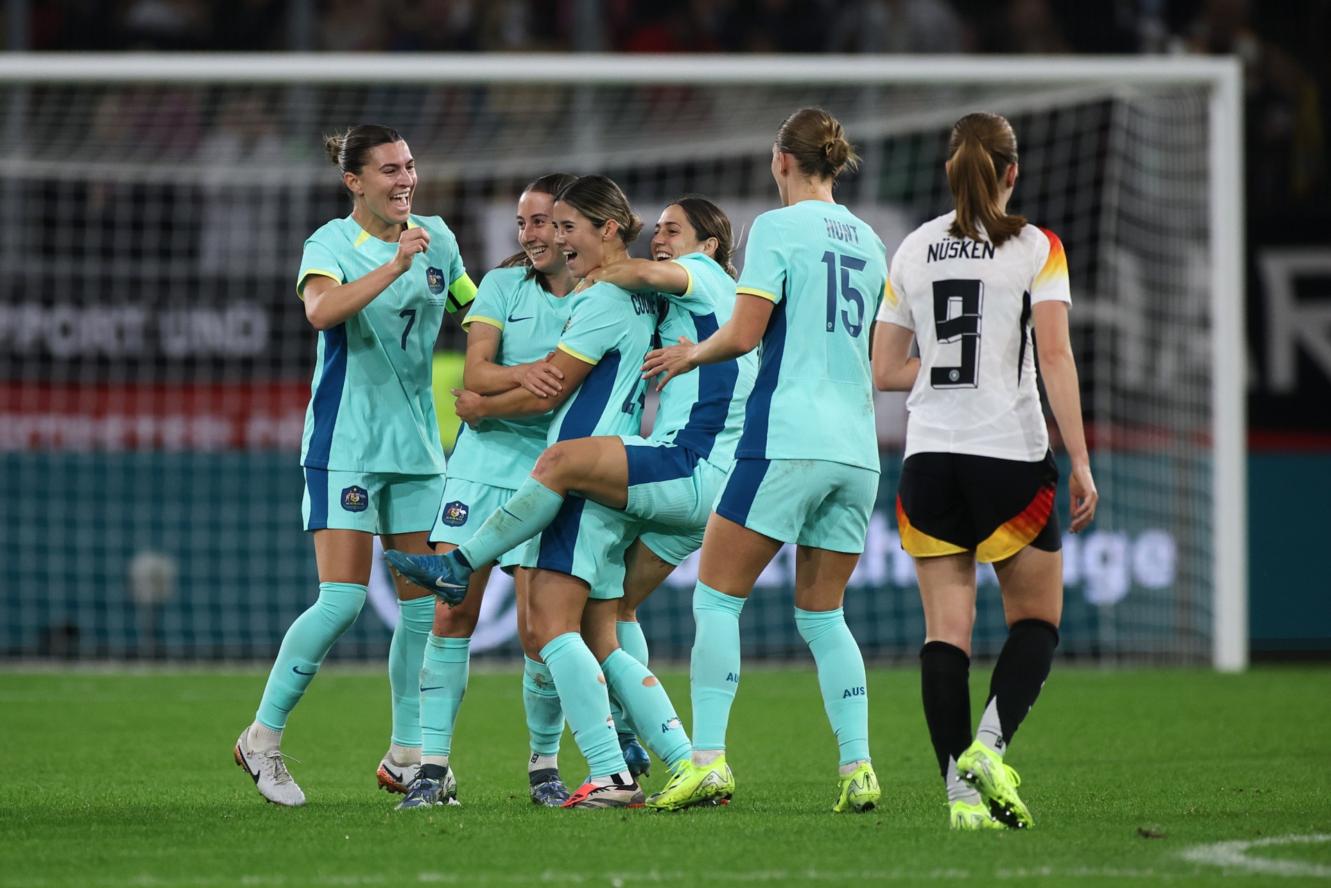 [{"clang_id":1,"value":"Frauenfußballspiel: Spielerinnen in türkisfarbenen und roten Trikots feiern ein Tor vor dem Tornetz. Rasen im Hintergrund."}]