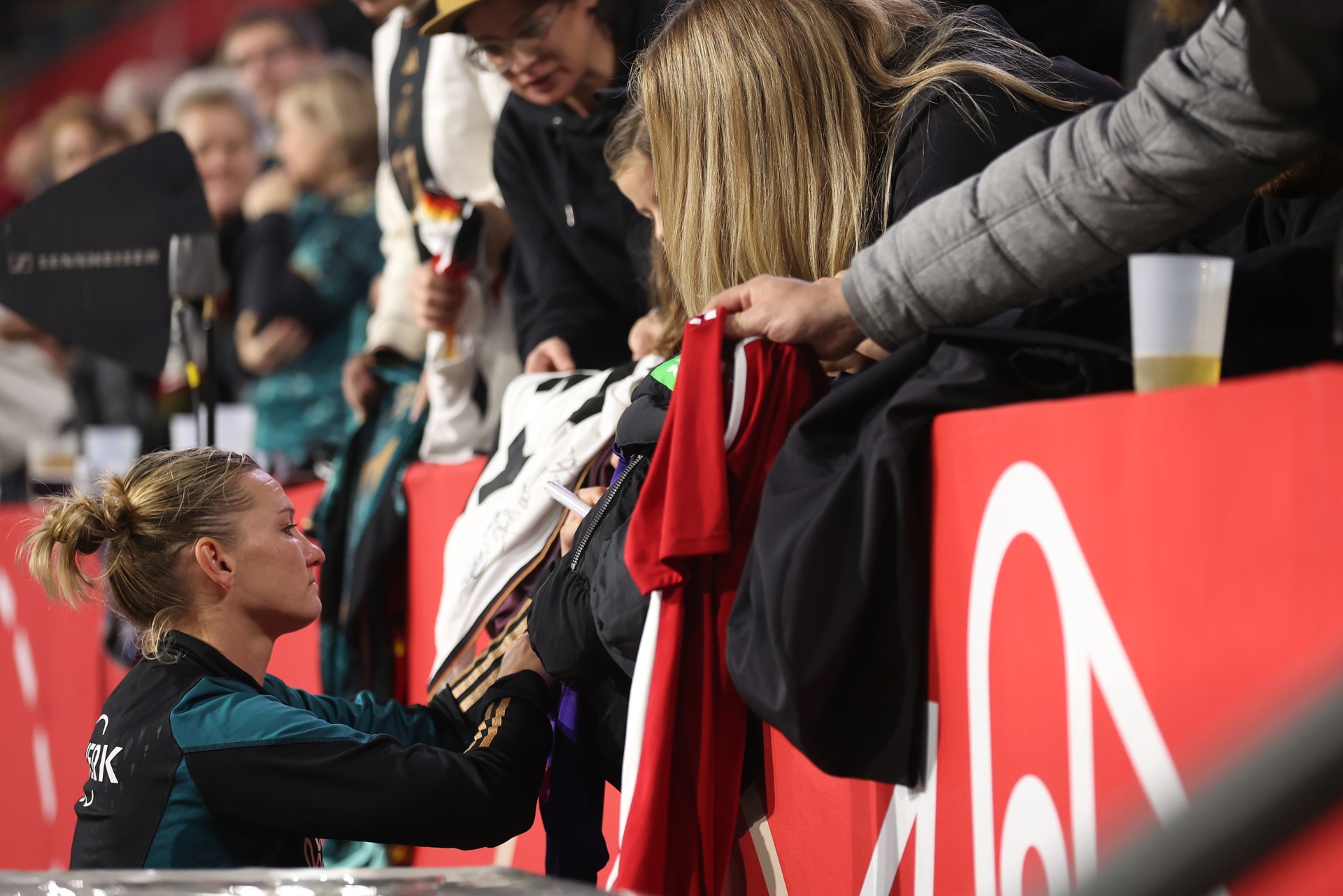[{"clang_id":1,"value":"Sportlerin in Trainingsjacke signiert Fan-Artikel über Barriere. Im Hintergrund Zuschauer und Werbebanner."}]