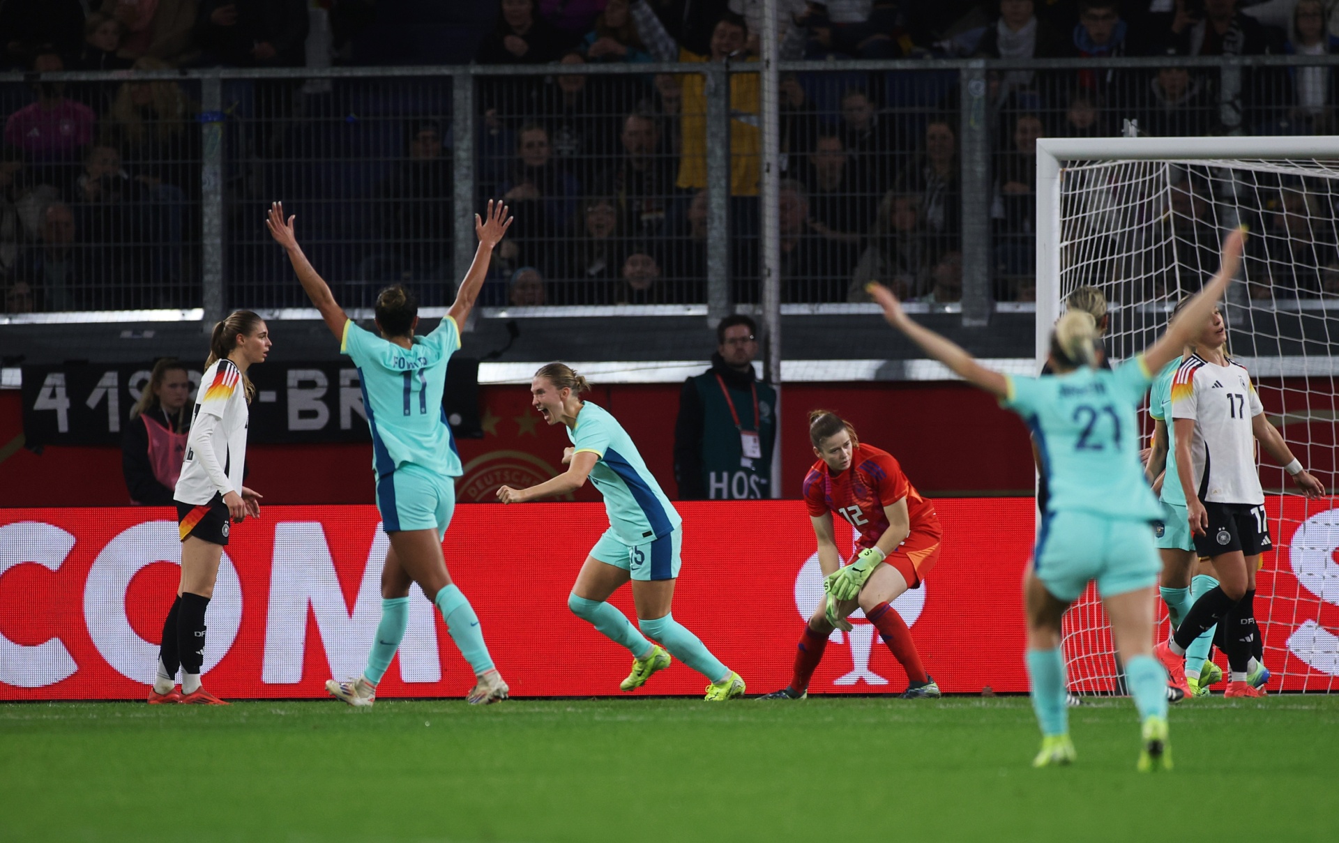 [{"clang_id":1,"value":"Frauenfußballspiel. Spielerinnen in türkisfarbenen und weißen Trikots jubeln mit erhobenen Armen vor dem Tor. Im Hintergrund Zuschauer und Werbebanner."}]
