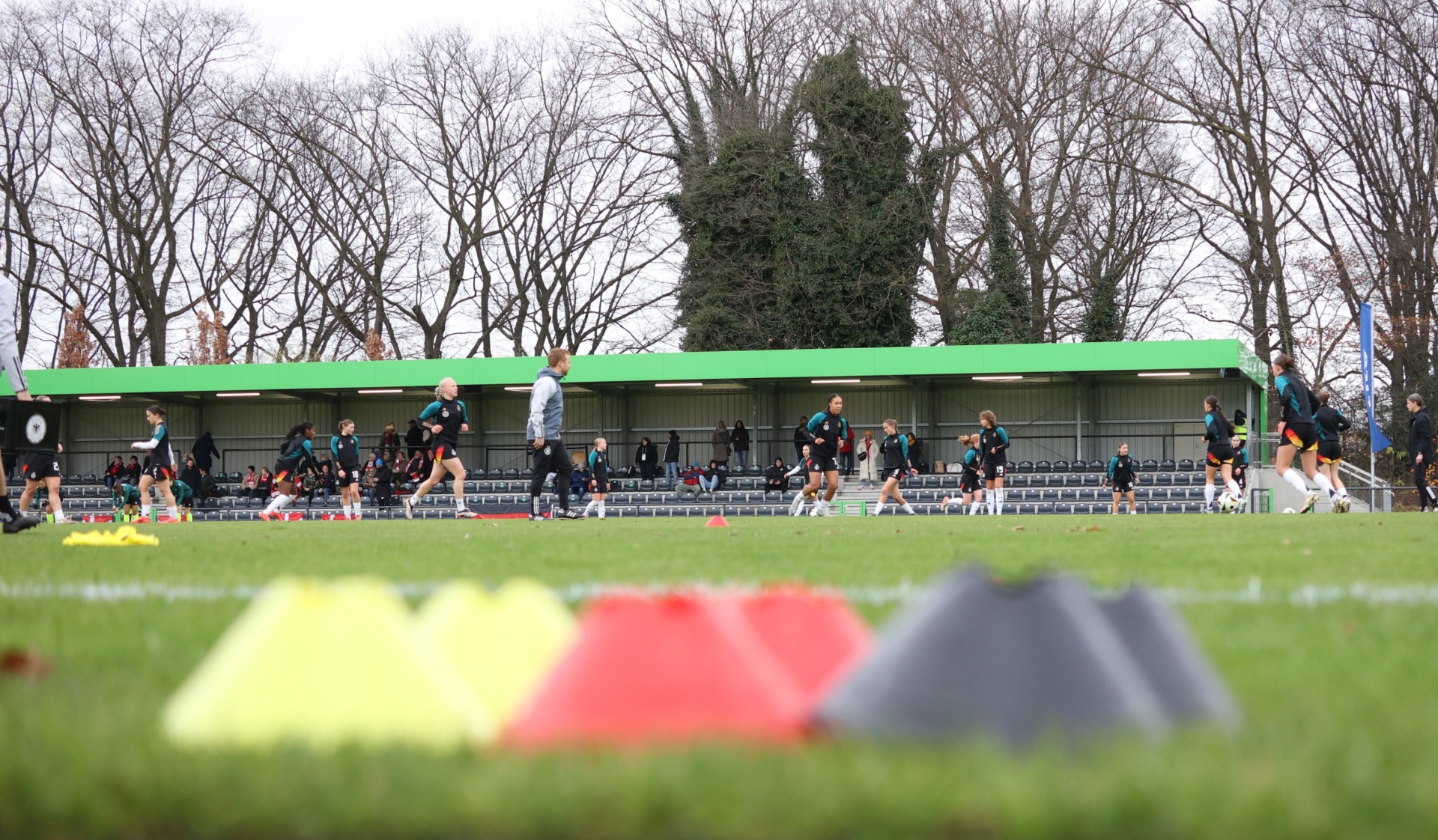 [{"clang_id":1,"value":"Fußballfeld mit Mannschaftsbank, Spieler im Trainingsbetrieb. Im Hintergrund Bäume ohne Blätter. Deutsche Flagge (Schwarz, Rot, Gelb) im Vordergrund."}]
