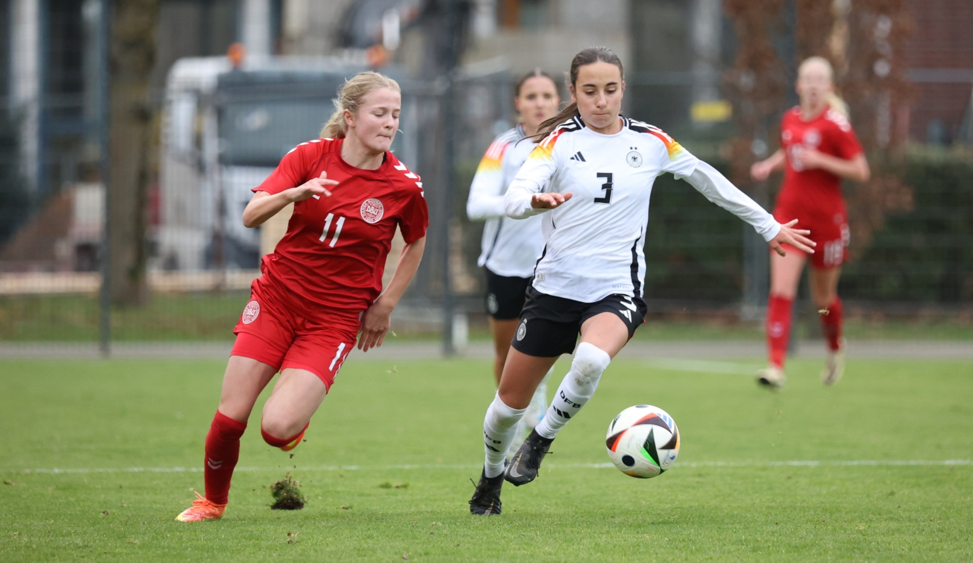 [{"clang_id":1,"value":"Fußballspiel: Spielerin mit Nummer 11 (Dänemark, rot) dribbelt gegen Spielerin mit Nummer 3 (Deutschland, schwarz-weiß) am Ball. Grasplatz, weitere Spieler im Hintergrund."}]