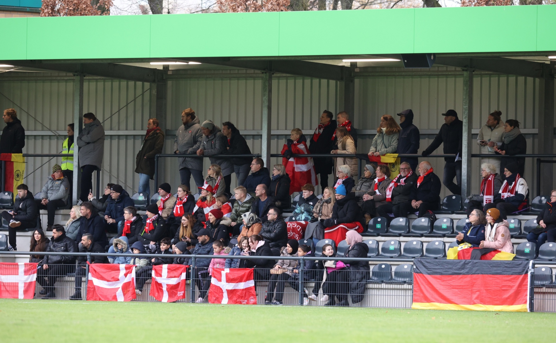 [{"clang_id":1,"value":"Fußballstadion: Zuschauerreihen mit Fans. Viele tragen rote Kleidung und schwenken dänische und deutsche Flaggen. Rasenfläche im Vordergrund."}]