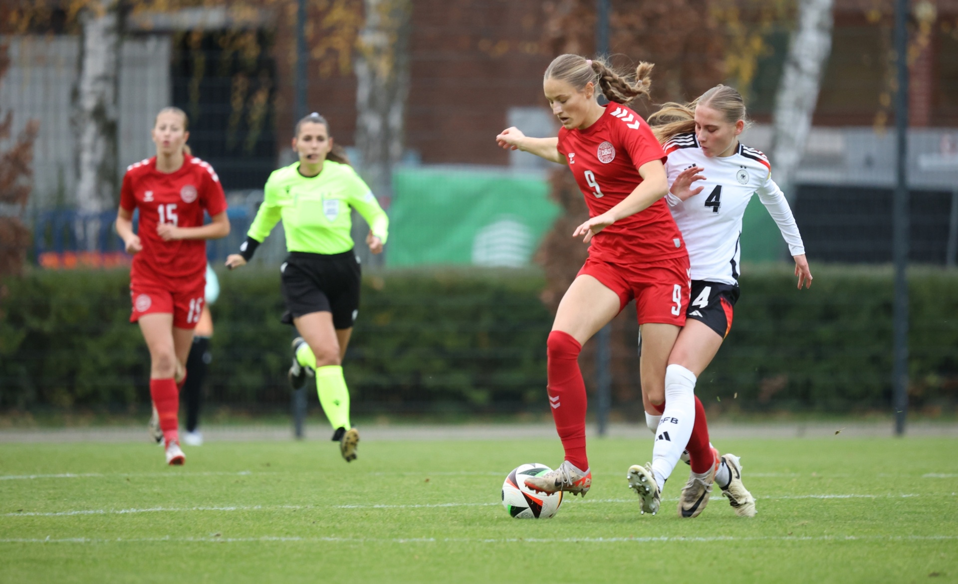 [{"clang_id":1,"value":"Zwei Fußballspielerinnen im Wettkampf. Links rotes Trikot mit Nr. 9, rechts schwarzes Trikot mit Nr. 4. Im Hintergrund weitere Spielerinnen und Bäume."}]