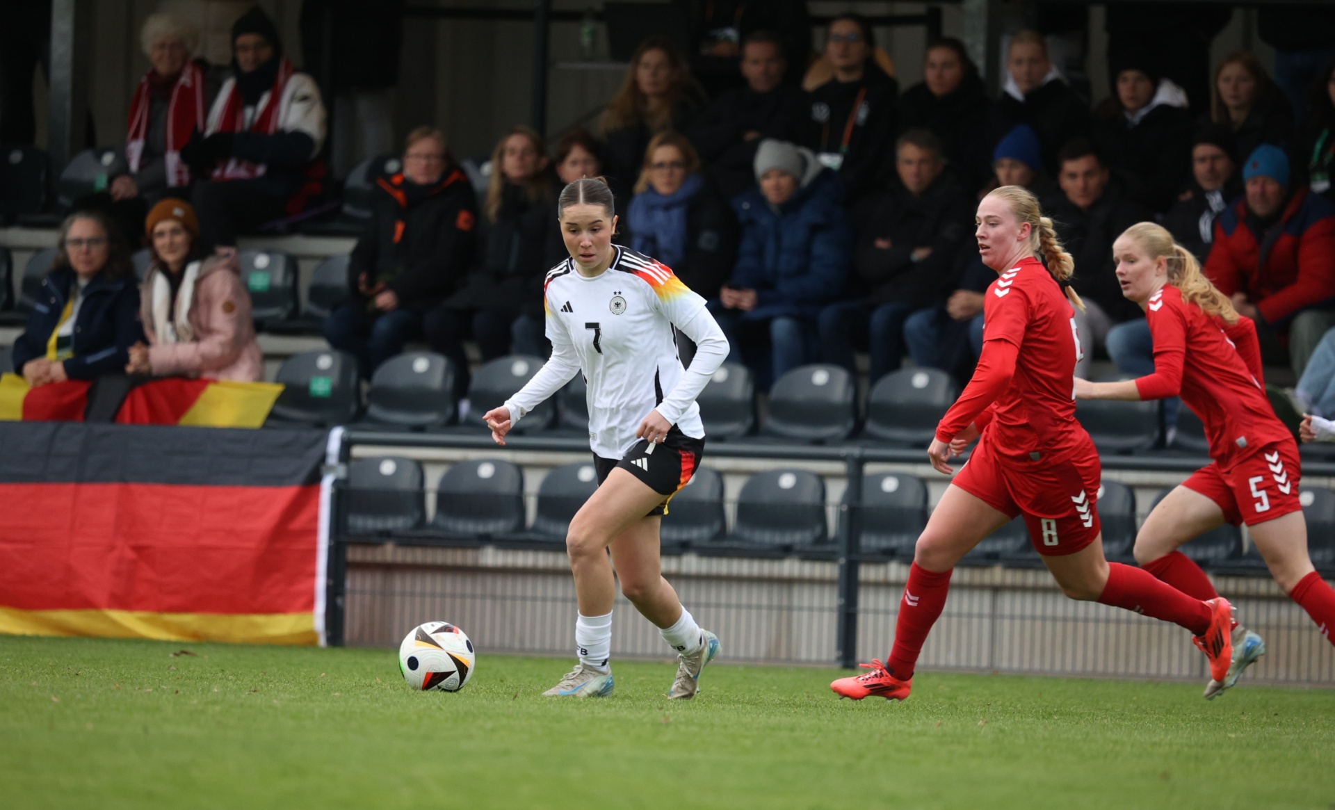 [{"clang_id":1,"value":"Fußballspiel: Spielerin mit Nummer 7 im weißen Deutschlandtrikot dribbelt den Ball. Zwei Gegenspielerinnen in roten Trikots folgen. Stadion im Hintergrund."}]
