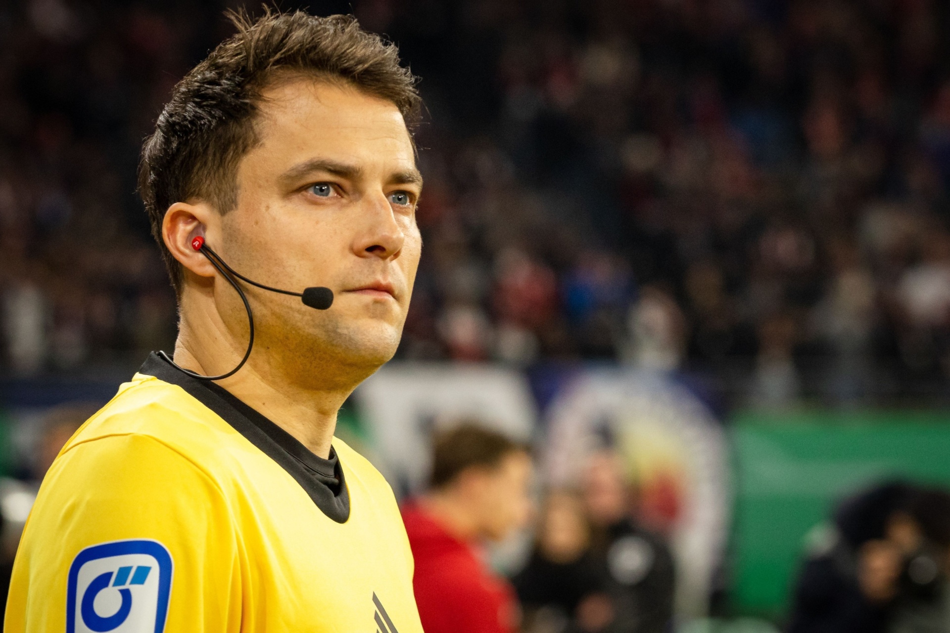 [{"clang_id":1,"value":"Fußballschiedsrichter mit Headset und gelbem Trikot vor verschwommenem Publikum und Spielfeld. Auf dem Ärmel ein Logo mit blauer Schrift auf weißem Grund."}]