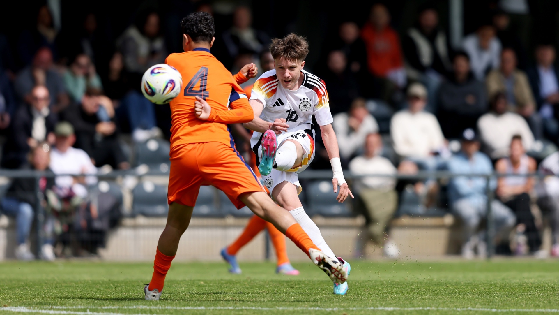 [{"clang_id":1,"value":"Zwei Fußballspieler im Spiel. Links in Orange, Nummer 4, rechts in Deutschlandtrikot, Nummer 10, mit Ball am Fuß. Stadion im Hintergrund."}]