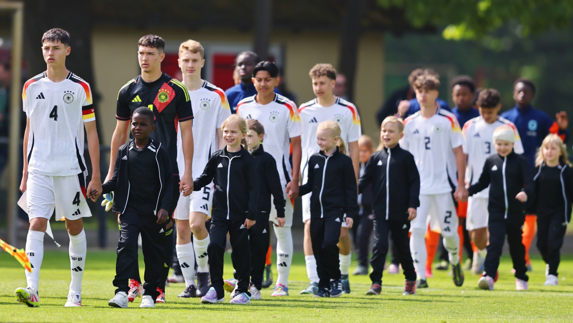 [{"clang_id":1,"value":"Fußballspieler in weißen Trikots mit schwarzen Akzenten und Kindern in schwarzen Jacken gehen auf einem grünen Rasenplatz. Im Hintergrund Gebäude."}]