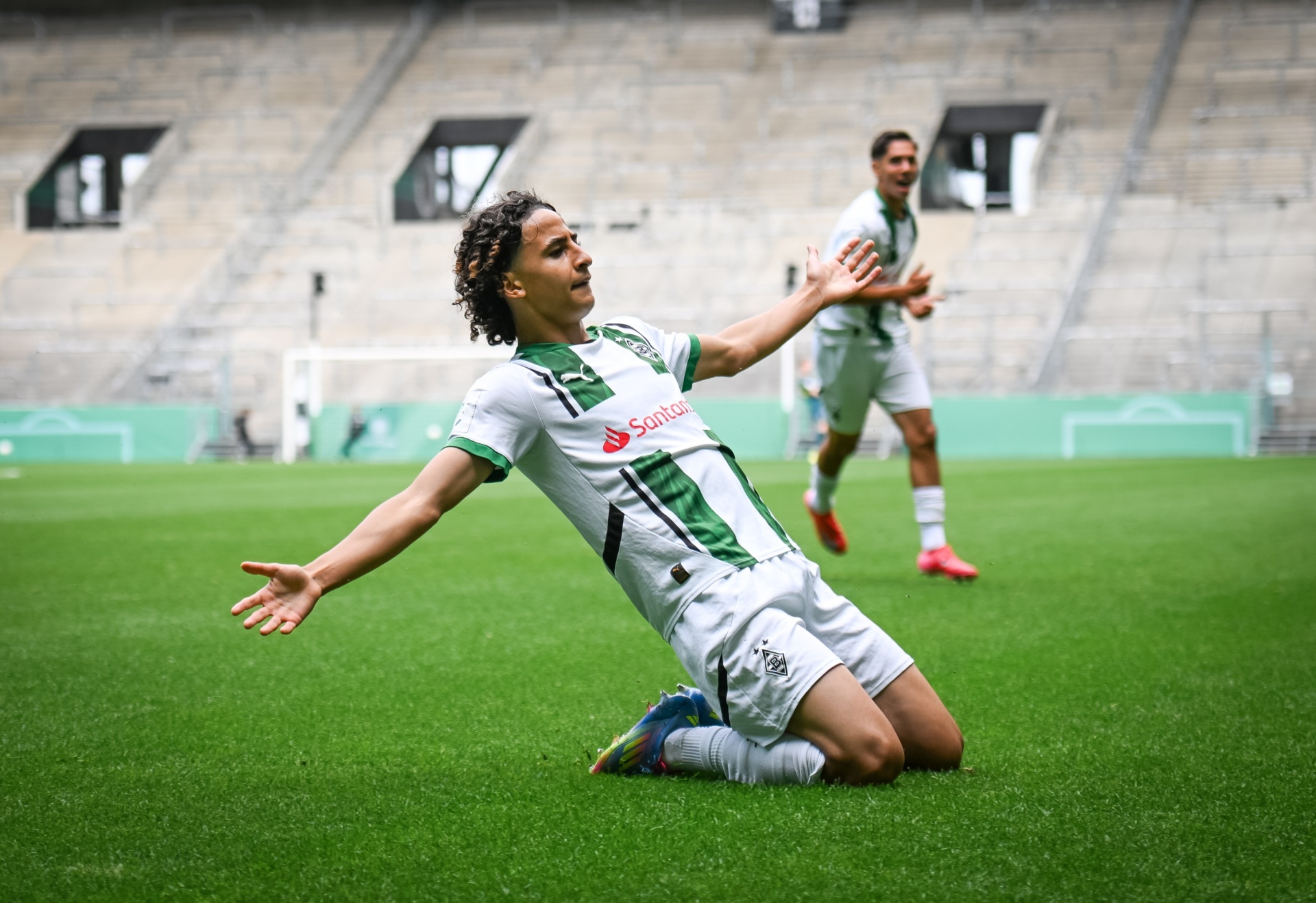 [{"clang_id":1,"value":"Fußballspieler kniet auf grünem Rasen, Arme weit geöffnet, jubelnd. Im Hintergrund ein leerer Stadionrang und ein weiterer Spieler."}]