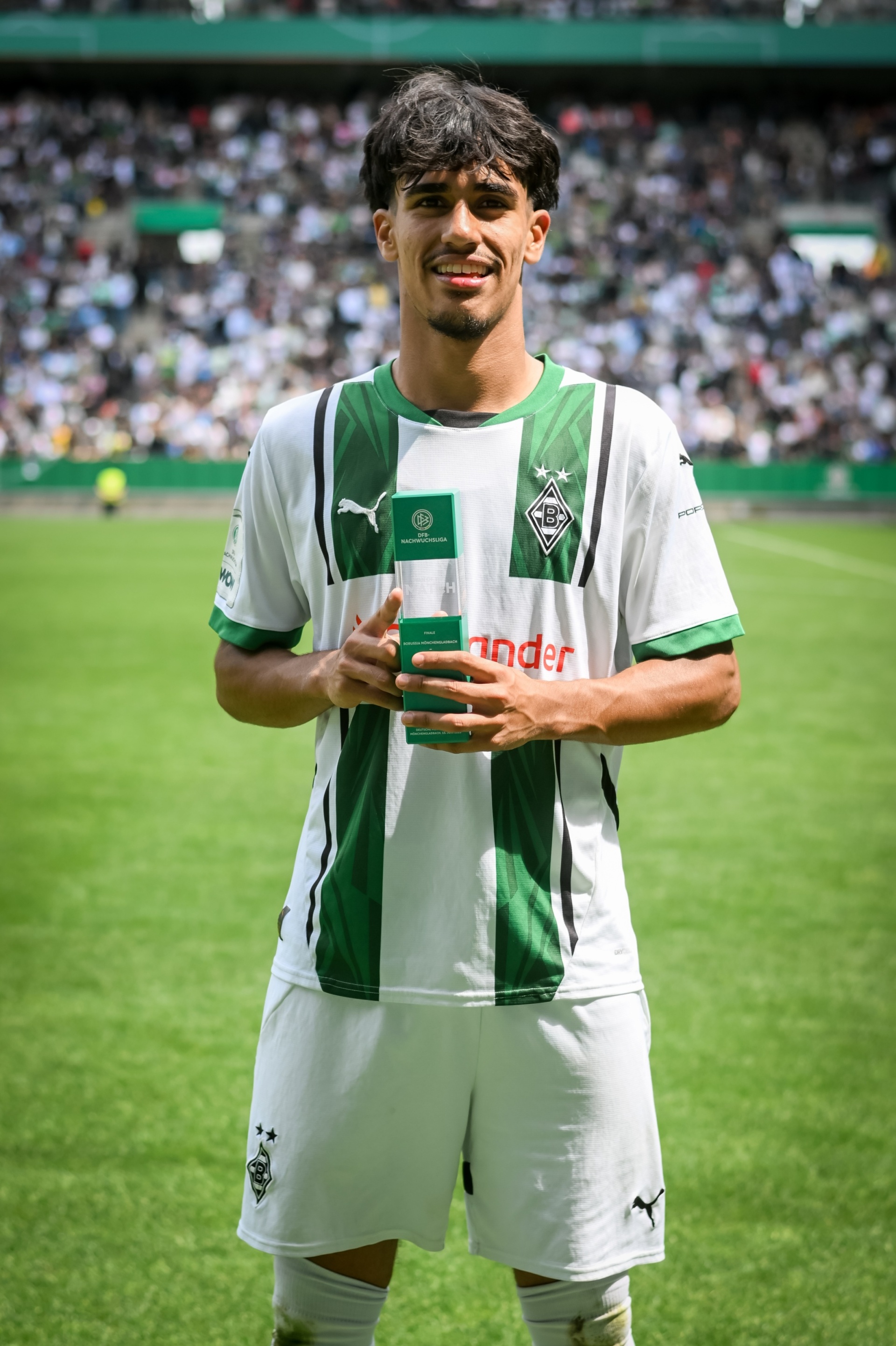 [{"clang_id":1,"value":"Fußballspieler mit dunklem Haar in grün-weißem Trikot und weißen Shorts steht auf einem Fußballfeld vor Zuschauermassen."}]