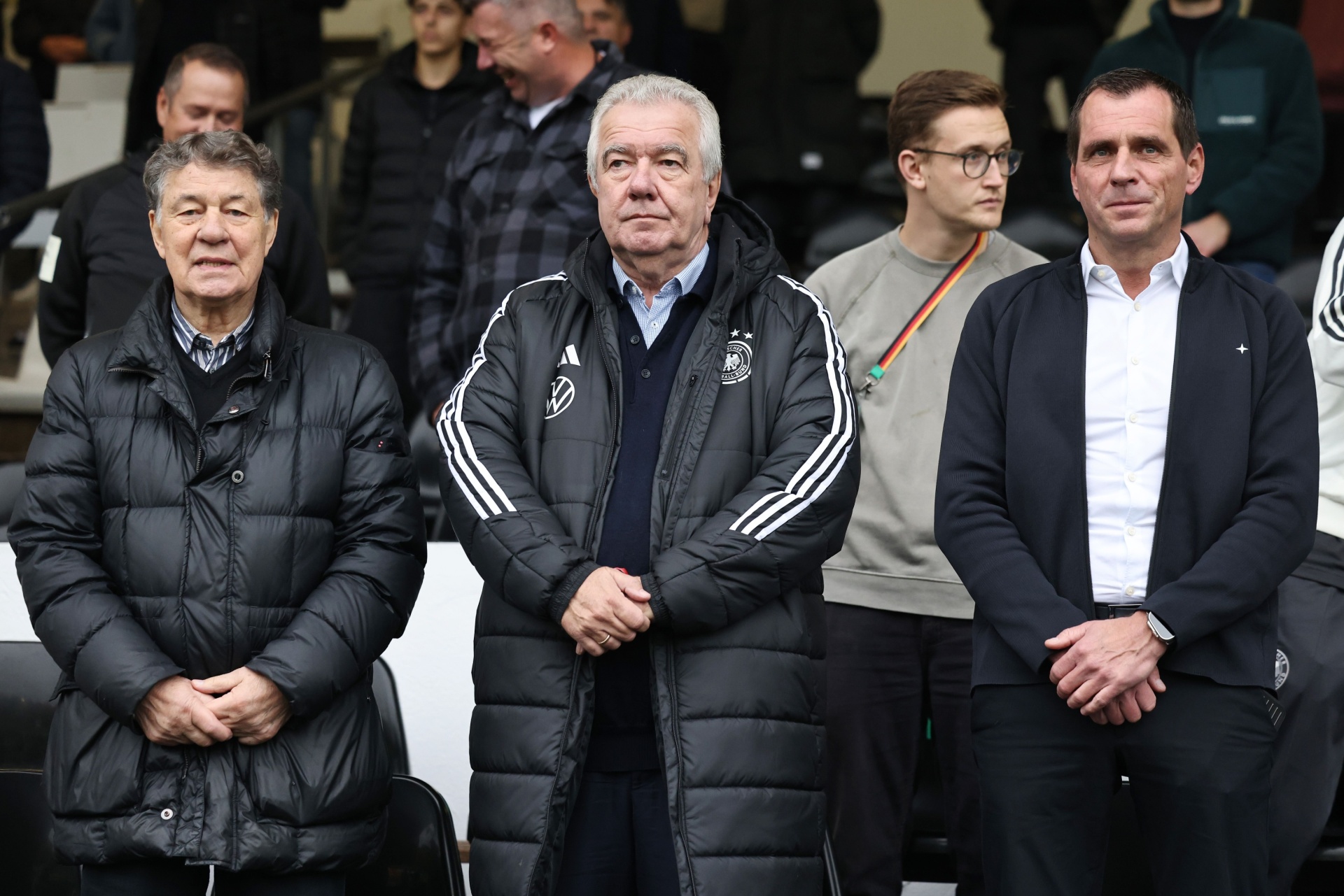 [{"clang_id":1,"value":"Vier Männer sitzen nebeneinander auf einer Tribüne. Sie tragen dunkle Jacken und schauen geradeaus. Im Hintergrund sind weitere Zuschauer zu sehen."}]